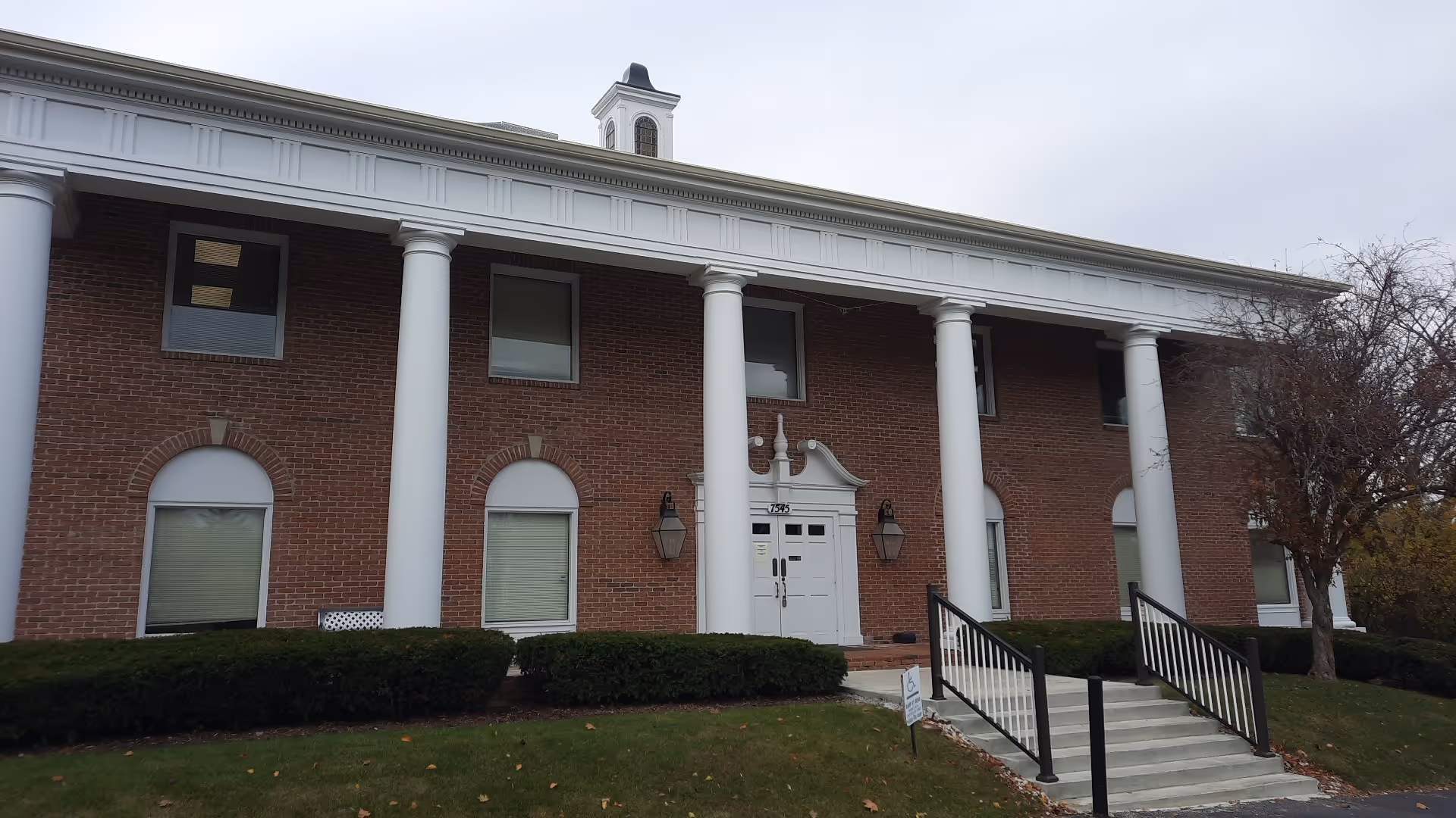 Brick building front with white columns and central entrance, surrounded by hedges.