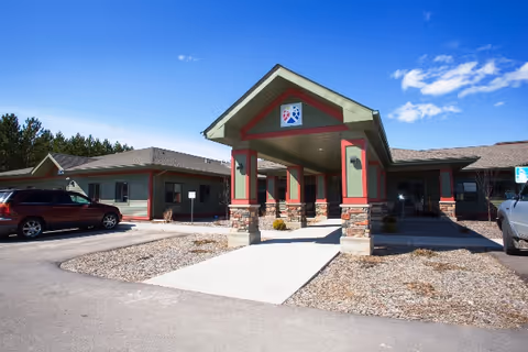 Exterior view of Carefree Living Babbitt facility showing the main entrance with a covered driveway supported by stone and wood pillars, surrounded by a parking area with cars and a clear blue sky.