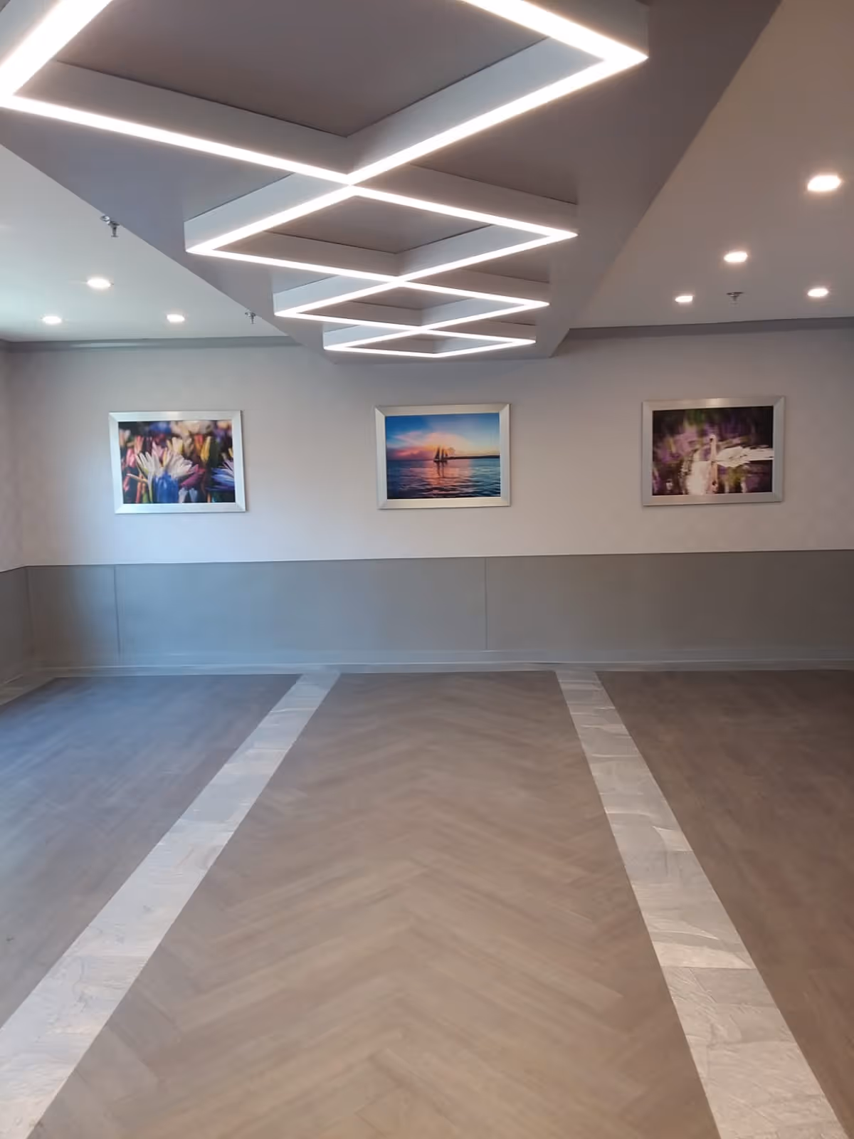 Empty interior common room with geometric ceiling lights, three framed pictures on the far wall, and patterned wood flooring.