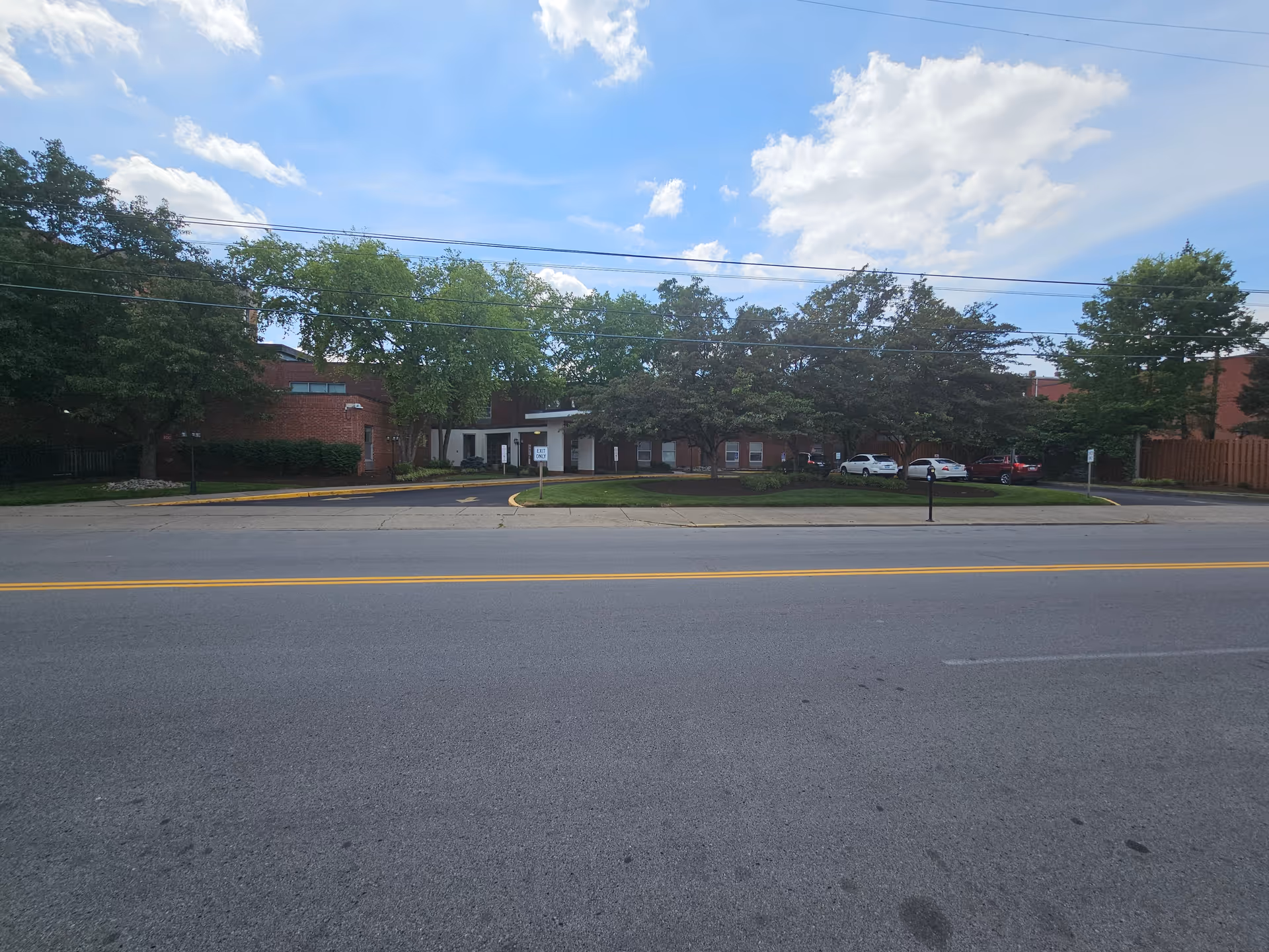 Front exterior of a brick senior living facility with trees, parked cars, and a street.