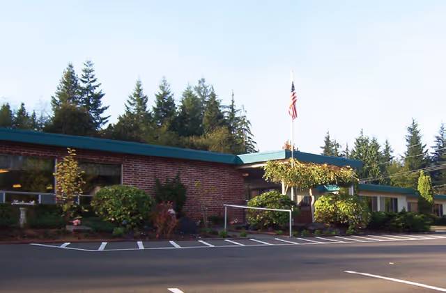 One-story brick building with a green roof, landscaped shrubs, an American flag, and parking spaces in front.