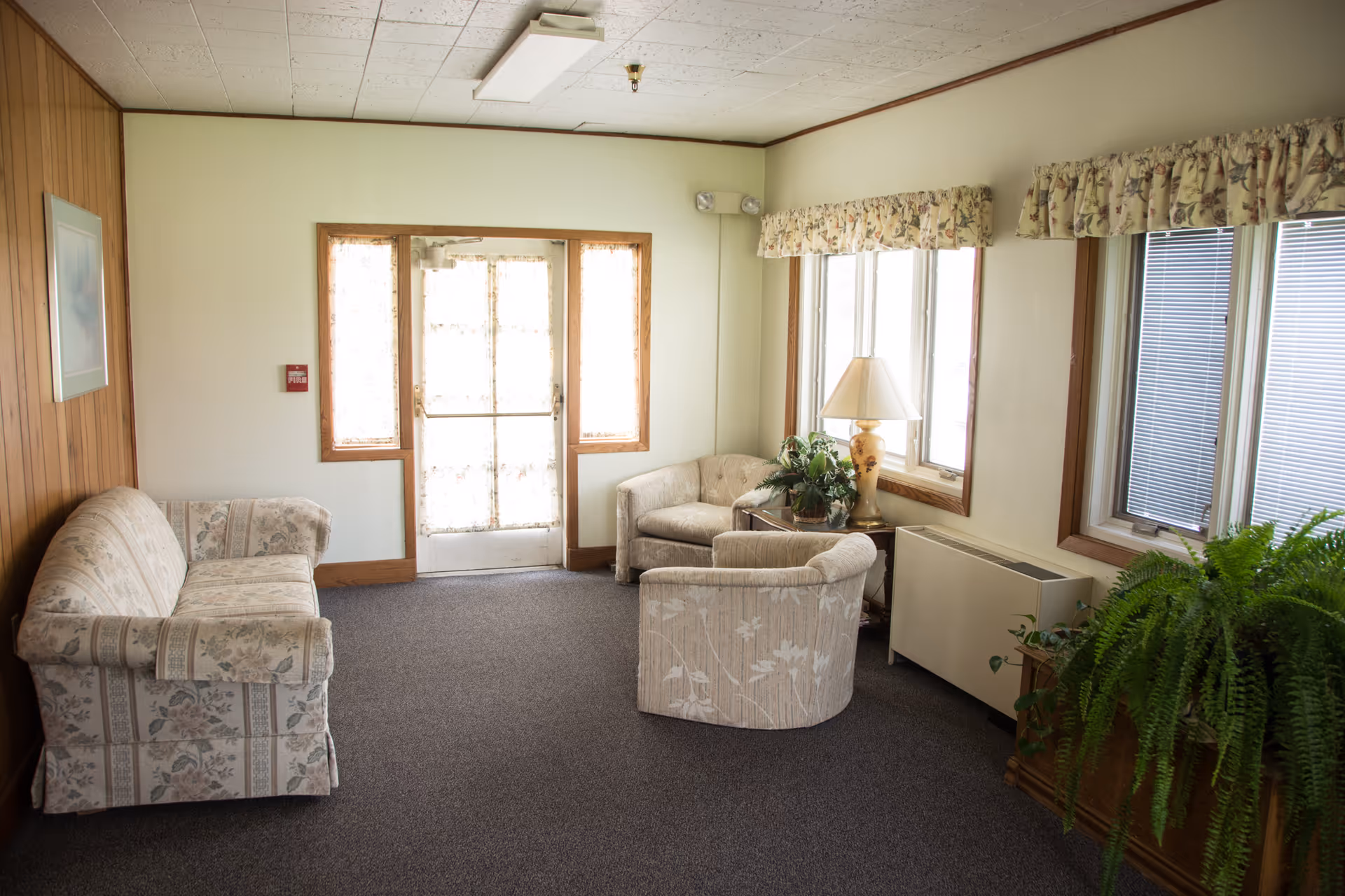 A small sitting area in an assisted living facility with a floral patterned sofa, two matching armchairs, a wooden side table with a lamp and a potted plant, windows with floral valances, and a door with sheer curtains allowing natural light to enter.