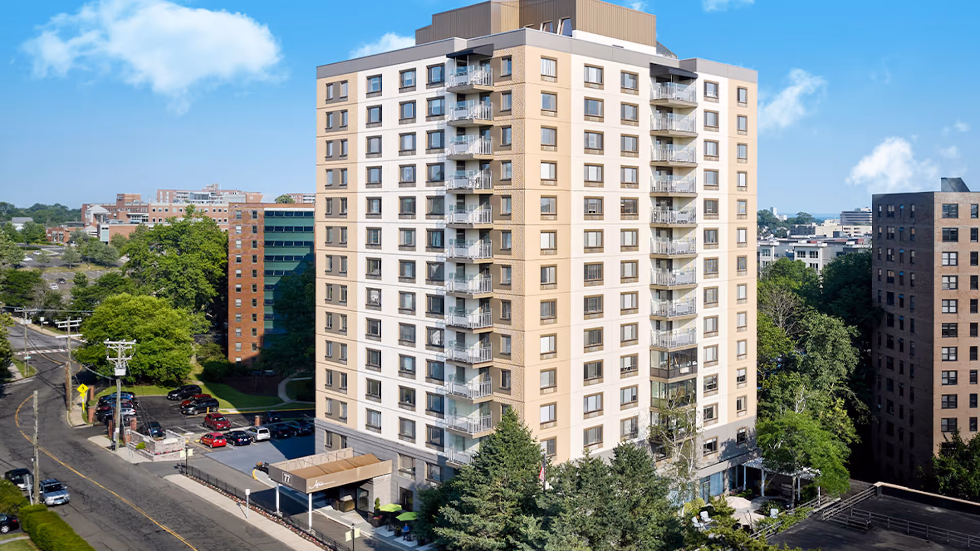 Tall beige senior living building with balconies, street, trees, and parked cars in view.