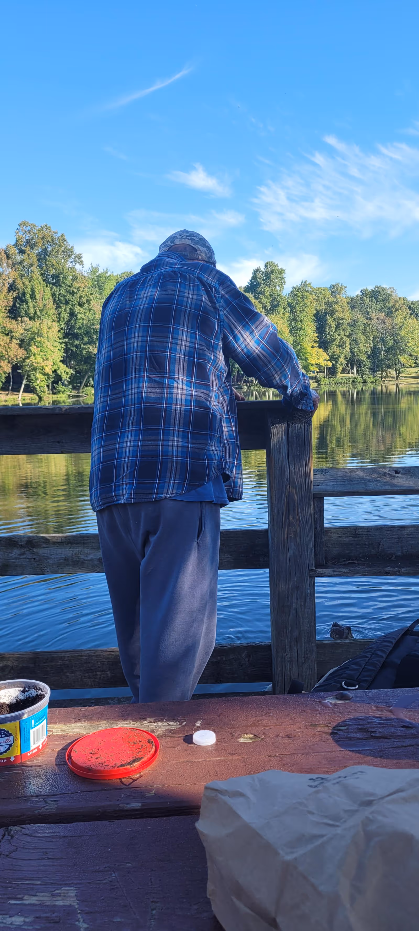 An older man in a blue plaid shirt stands at a wooden railing by a lake.
