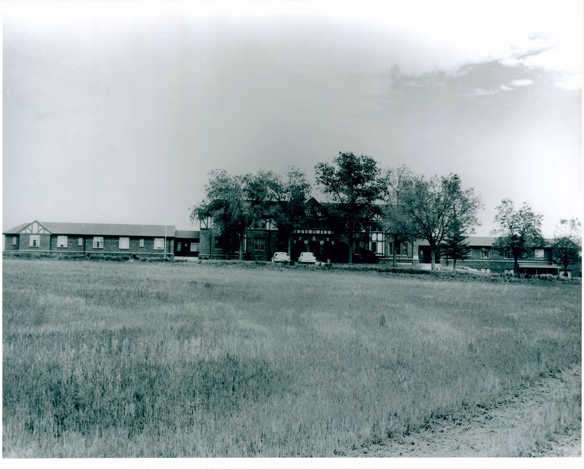 Black and white photo showing the front exterior view of a large building with multiple sections and a few trees in front. There are two vintage cars parked near the entrance, and an open grassy field extends in the foreground.