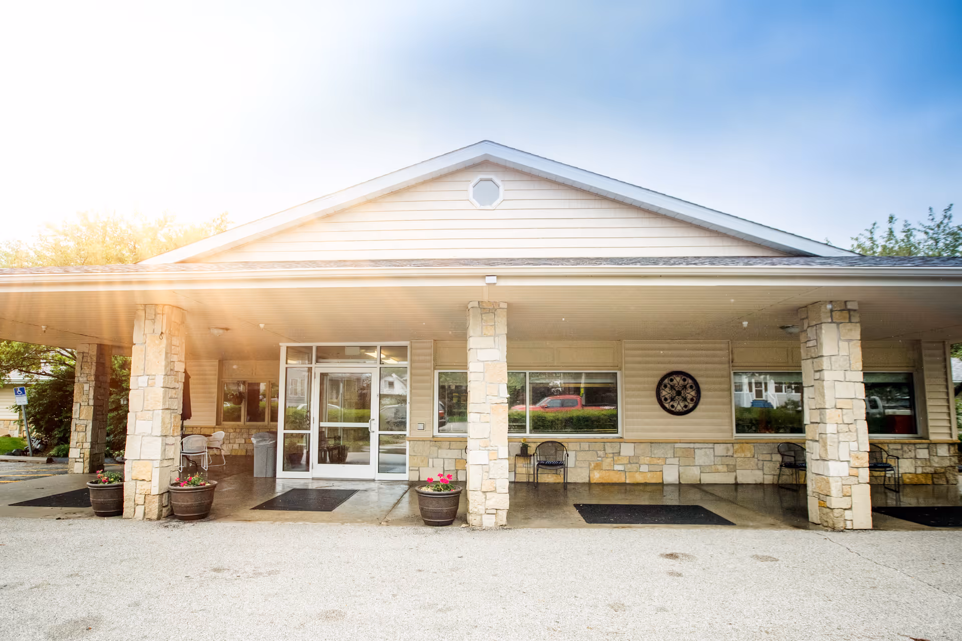 Front entrance of a senior living facility with a covered porte-cochère, stone columns, glass doors, planters, and outdoor seating.