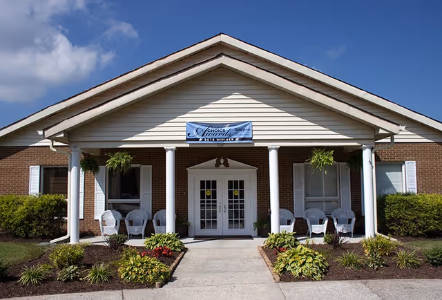Front exterior view of a single-story brick building with white columns supporting a peaked roof. There are white double doors at the entrance, flanked by windows with white shutters. Several white chairs are arranged on the porch, and there are hanging plants and landscaped bushes and flowers along the walkway leading to the entrance. A banner above the door reads 'Choice Awards 2012 Winner'.