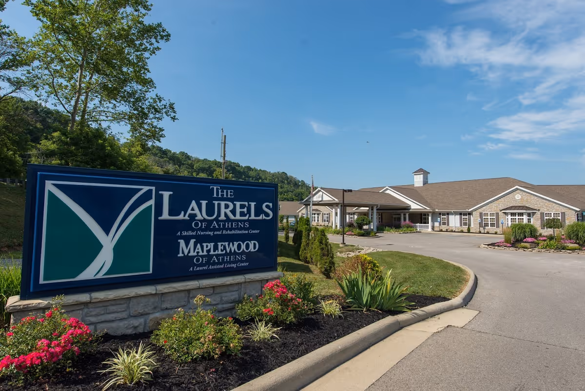 Exterior view of The Laurels of Athens, a skilled nursing and rehabilitation center, showing the facility building with a driveway and landscaped garden with flowers and shrubs under a clear blue sky.