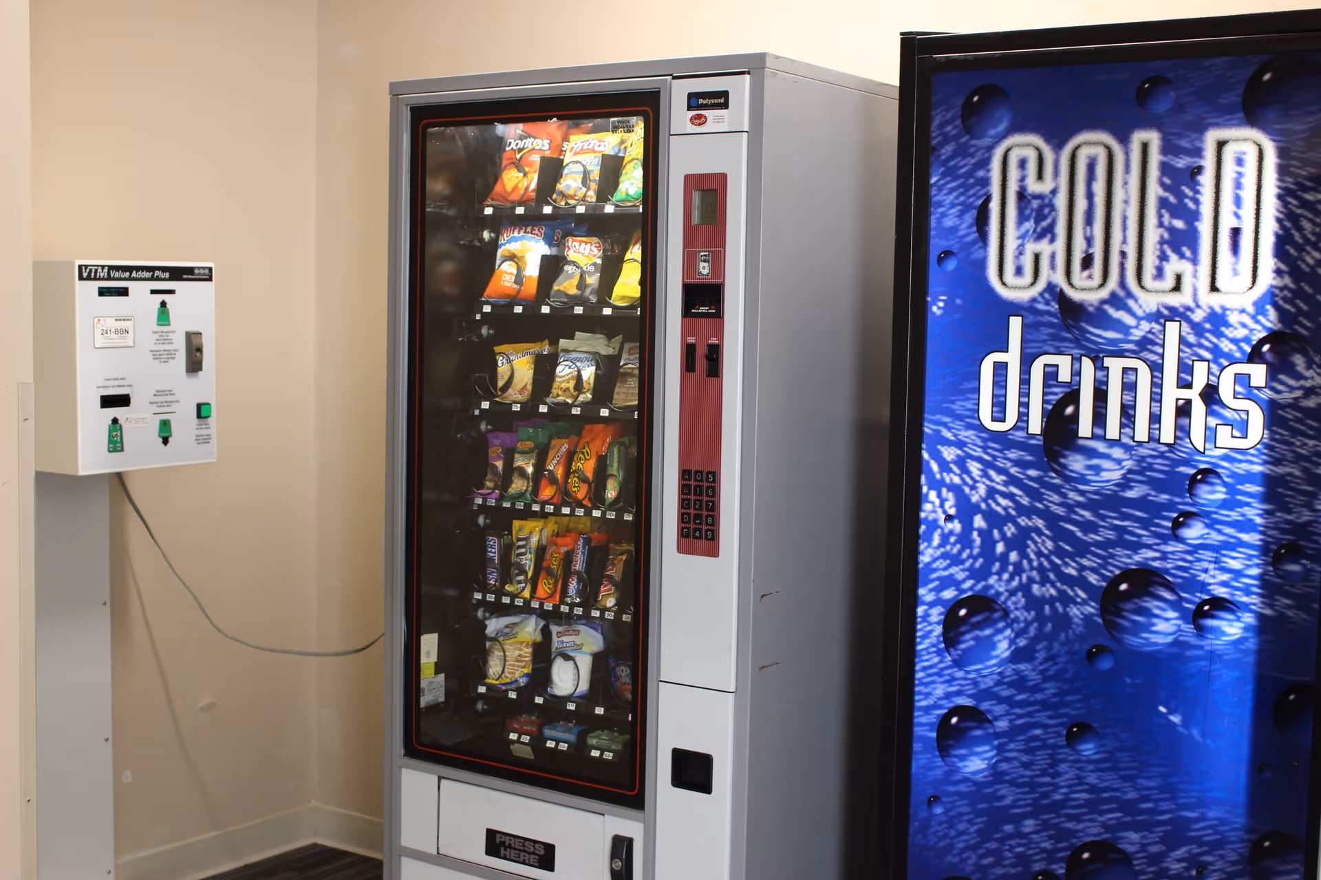 A snack vending machine next to a blue 'COLD drinks' vending machine against a beige wall.