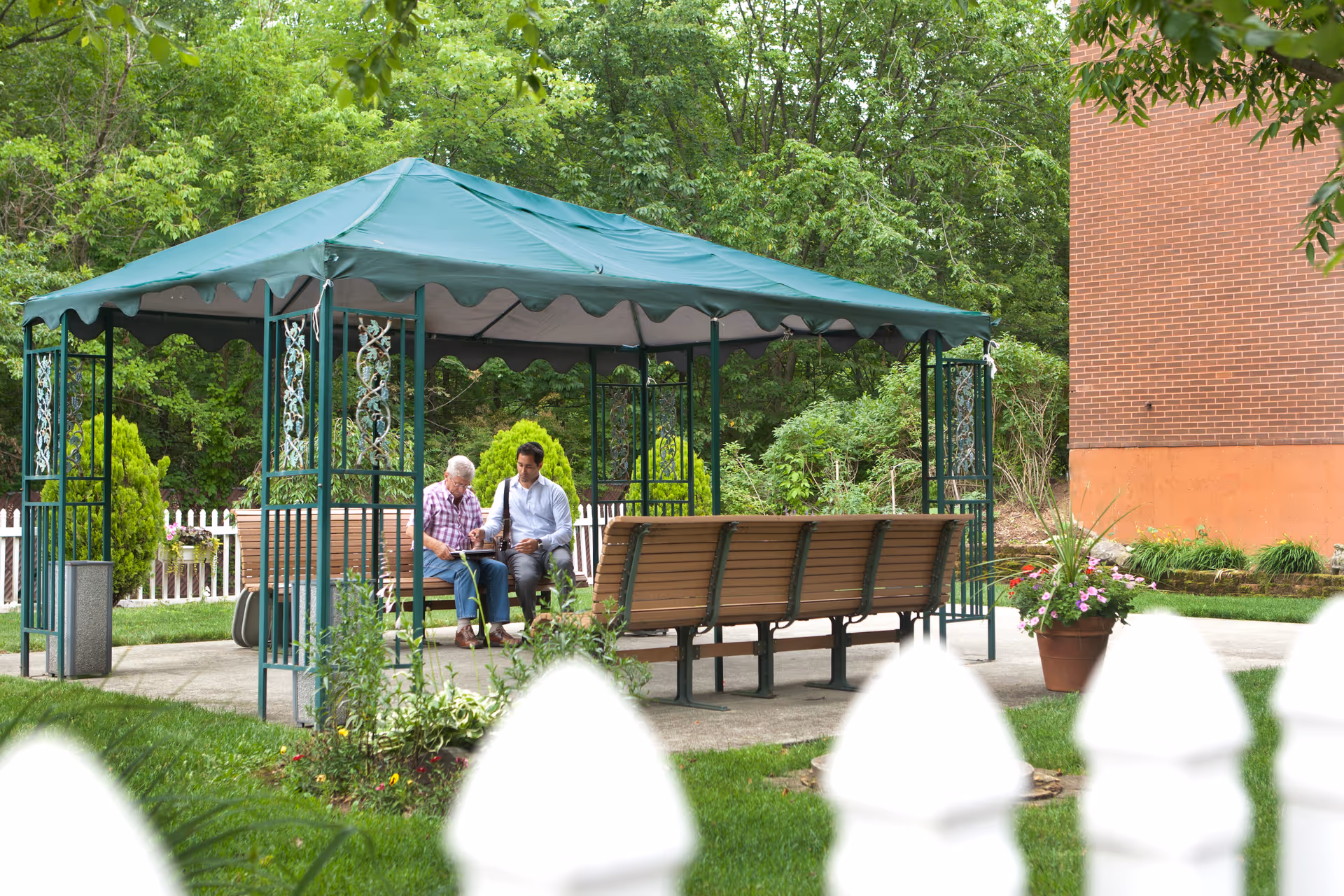 Two people sit and chat under a green gazebo in a garden near a brick building.