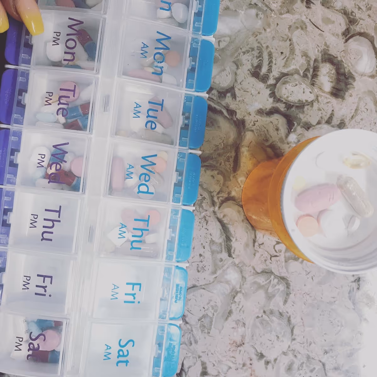 Weekly pill organizers labeled by day with assorted pills on a marble counter.