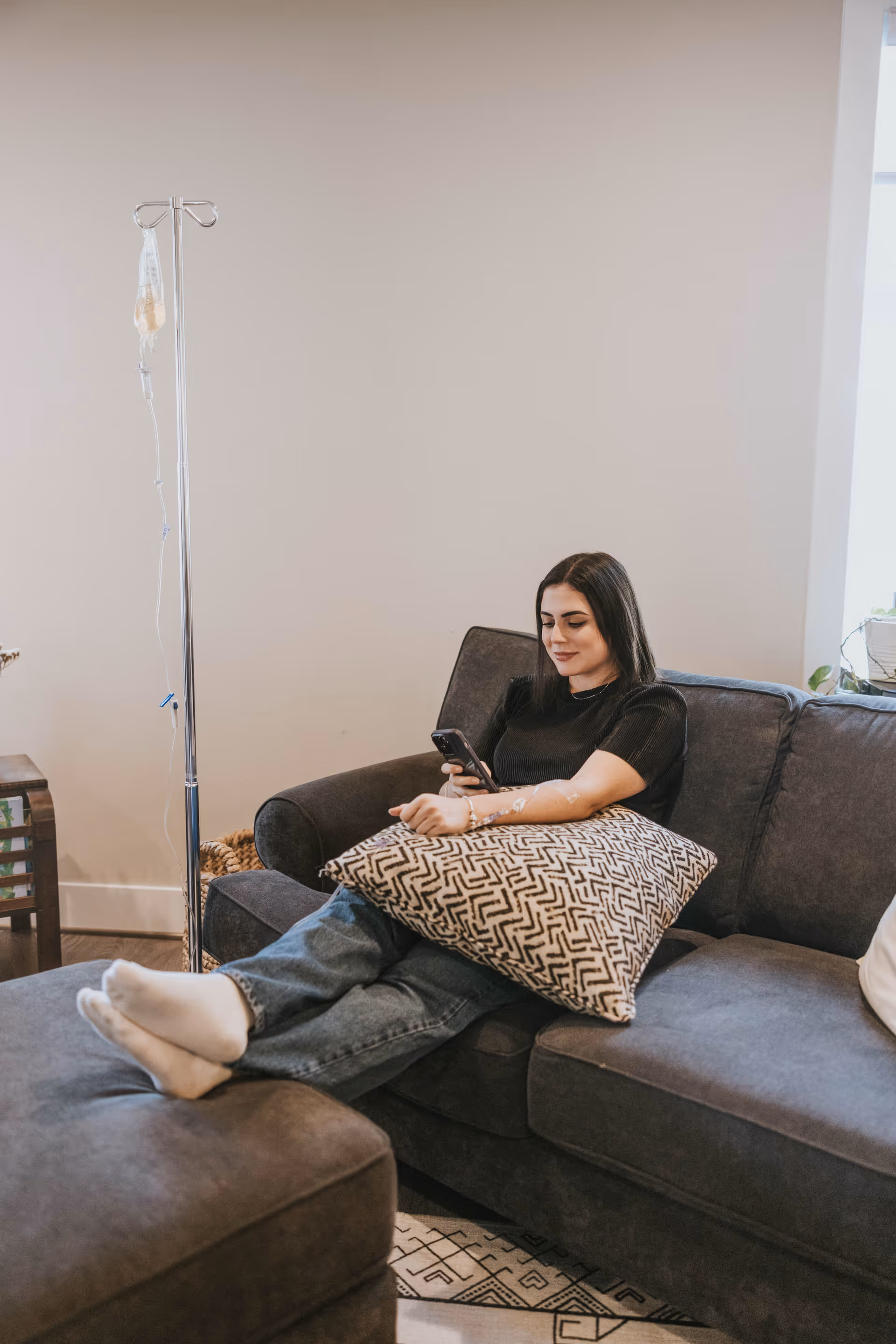 A woman lounges on a sofa in a modern living room beside an IV stand.