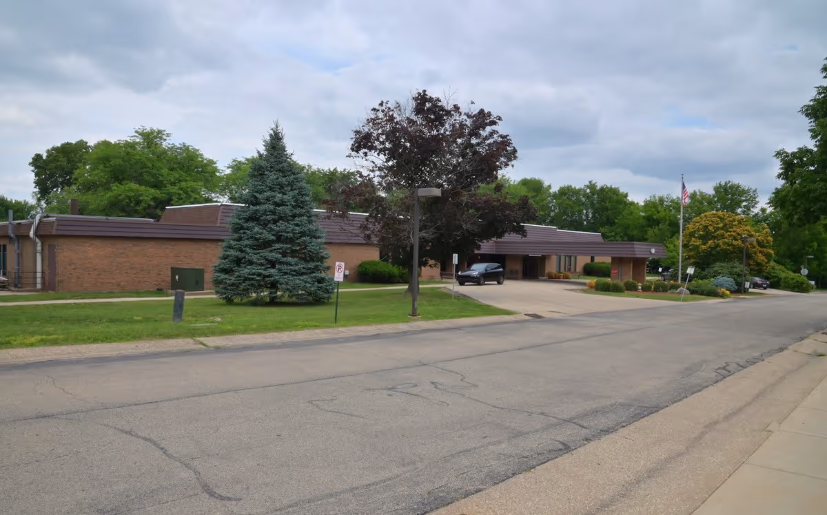 Exterior view of a single-story brick senior living facility with a driveway, trees, and parked cars.