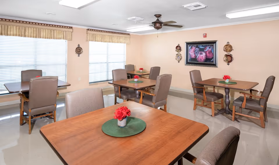 Bright communal dining room with several wooden tables and cushioned chairs, each table decorated with a small vase of red flowers.