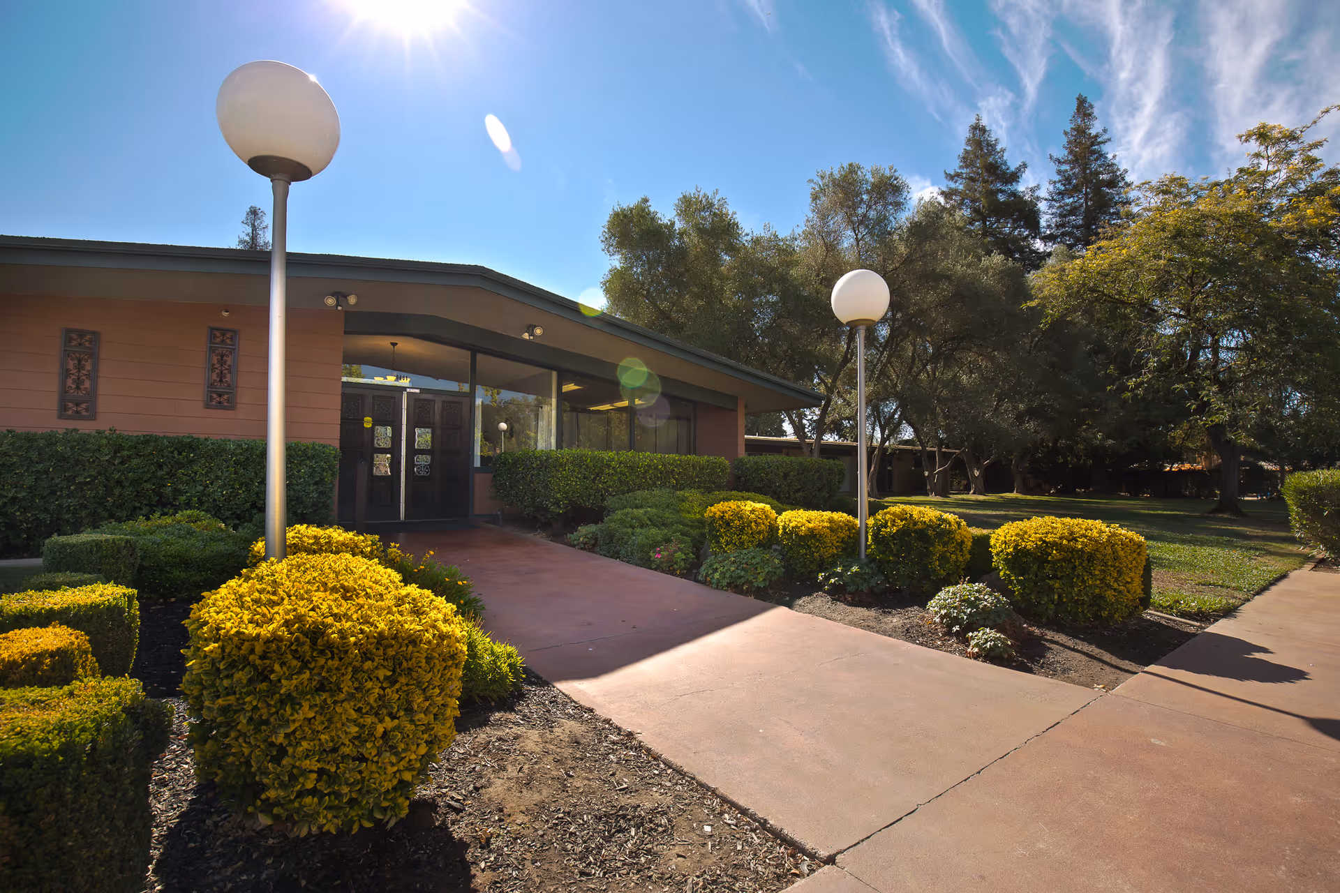 Entrance of a single-story care facility with a walkway, landscaped shrubs, and globe lamp posts under a sunny sky.