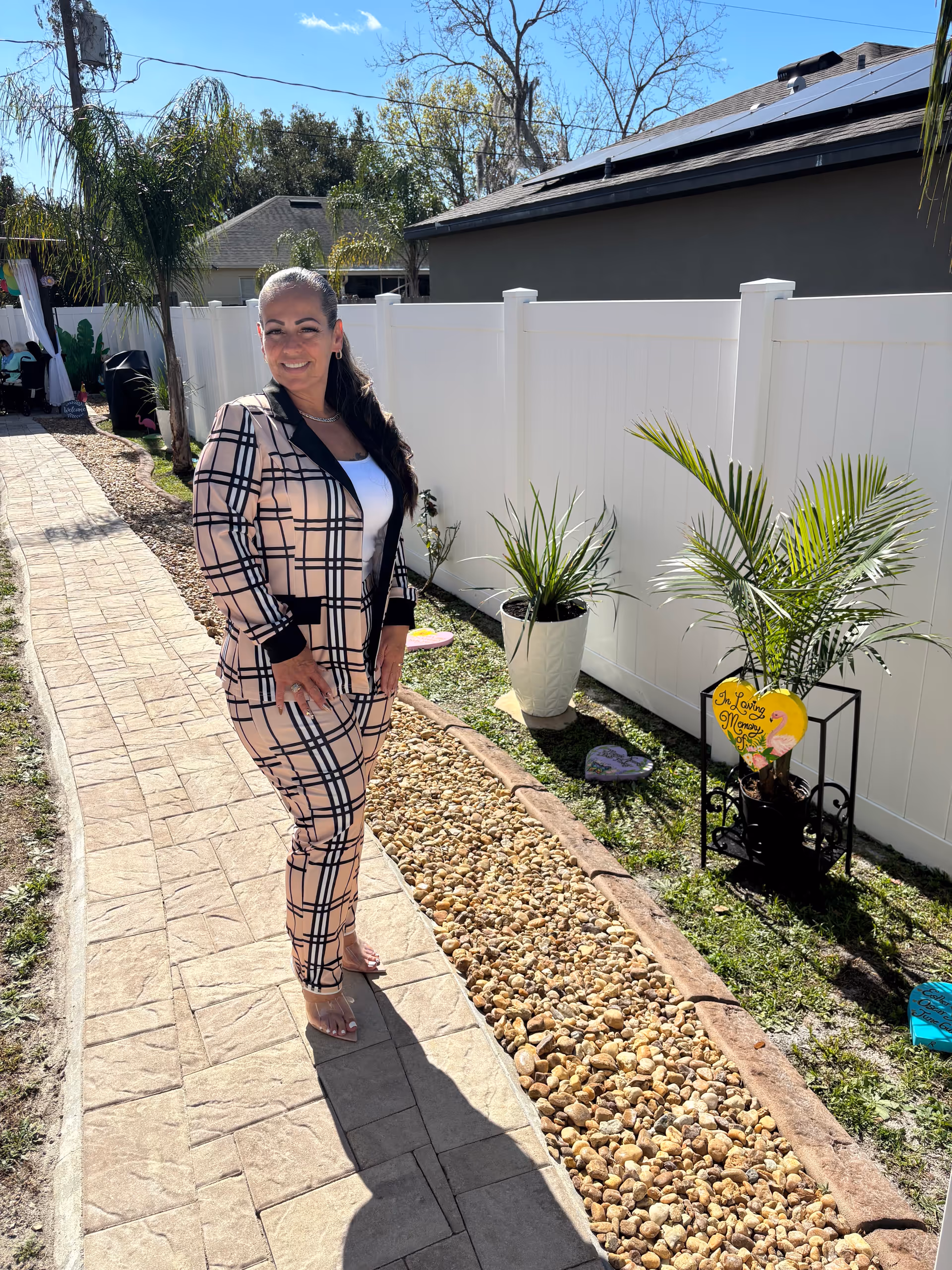 Smiling woman in plaid suit stands on a stone path beside a white fence outdoors