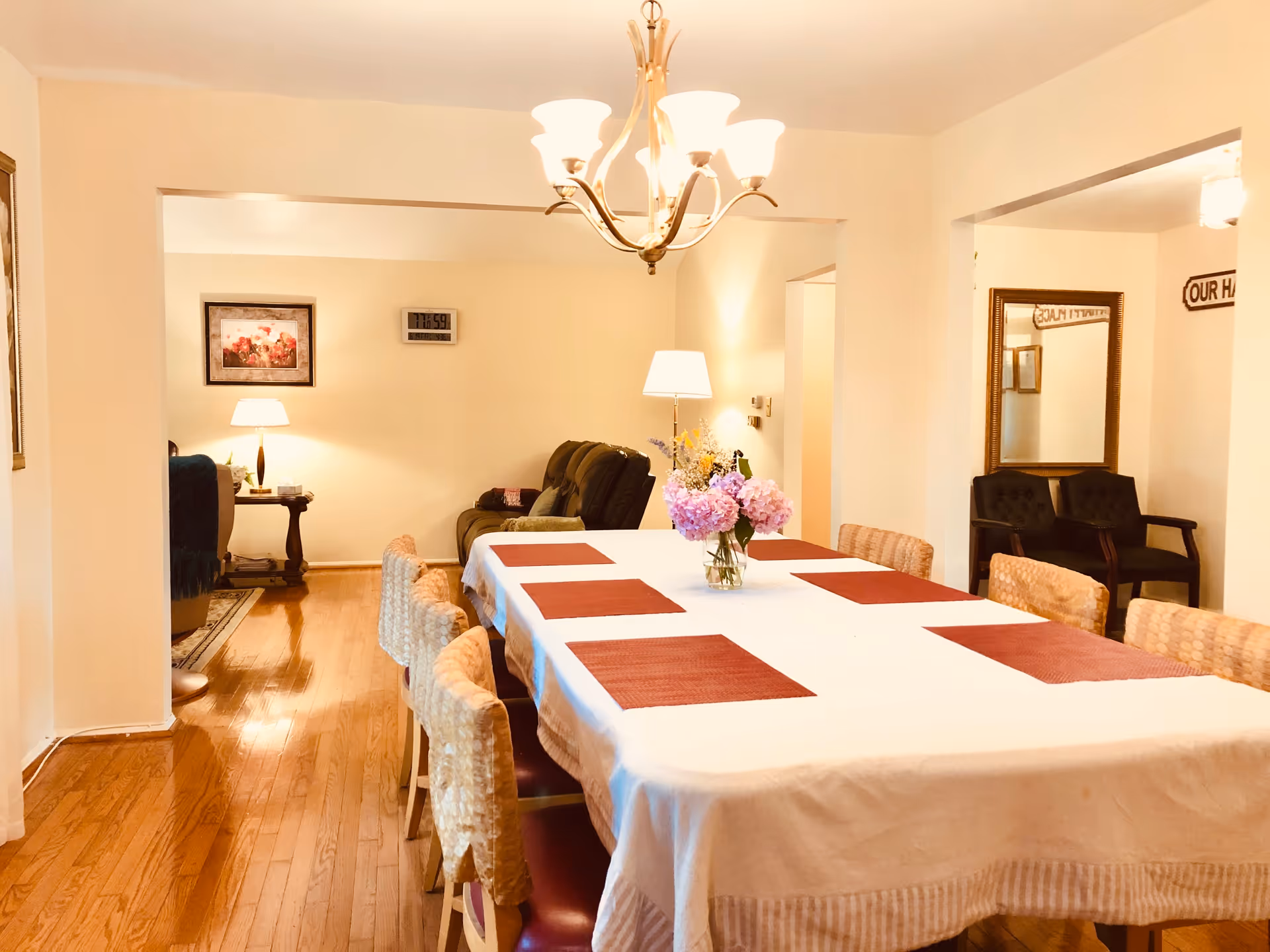 A dining room with a long table covered with a white tablecloth and red placemats. There is a vase with pink flowers in the center of the table. The room has wooden flooring, a chandelier hanging above the table, and a living area with a couch and lamps visible in the background. There is also a mirror and two chairs against the wall on the right side.