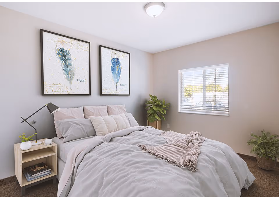 A cozy bedroom with a neatly made bed featuring light gray and pink bedding, a wooden nightstand with a modern black lamp, a small plant, and a cup. Two framed feather artworks hang above the bed. A window with white blinds lets in natural light, and there are two potted plants in the corners of the room.