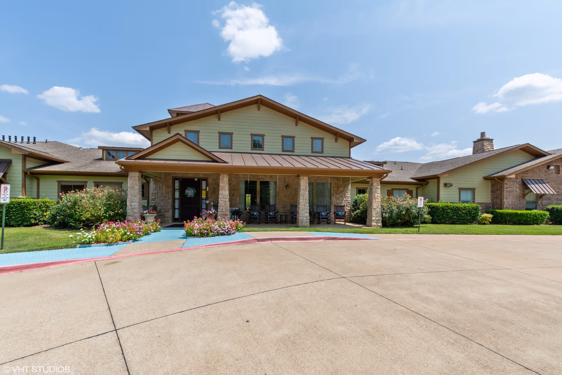 Front exterior view of Azalea Trails Assisted Living and Memory Care facility showing a single-story building with a covered entrance supported by stone pillars, surrounded by green bushes and colorful flowers under a partly cloudy blue sky.