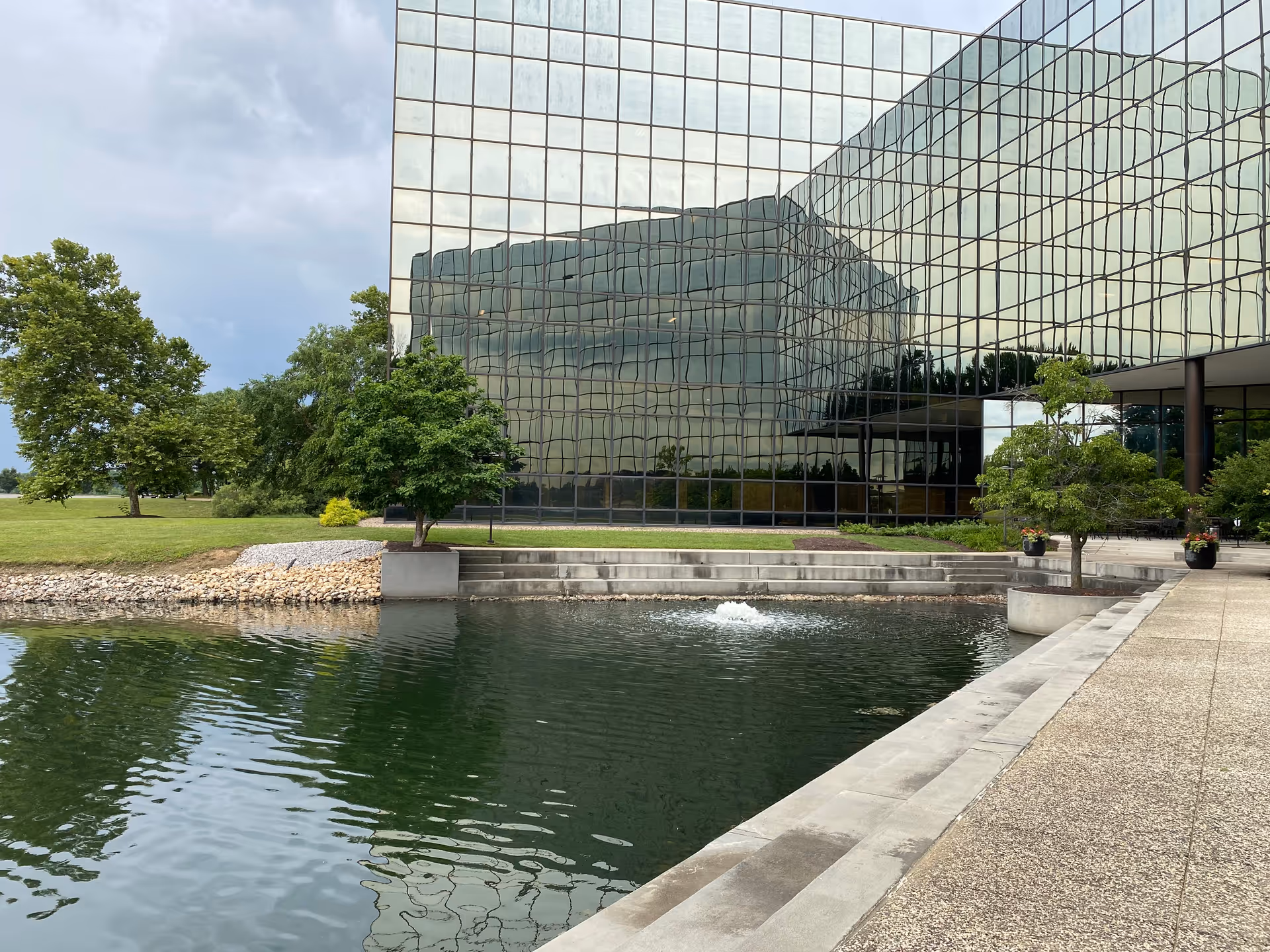 Modern glass-front building with a reflective exterior, water feature, trees, and a wide paved walkway.