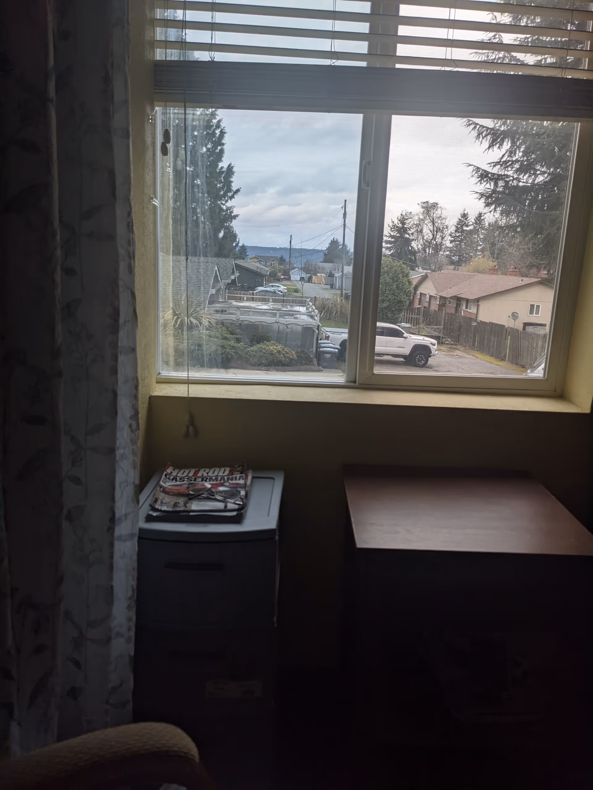 Dim room with a window, blinds, small cabinet, and a magazine on top.