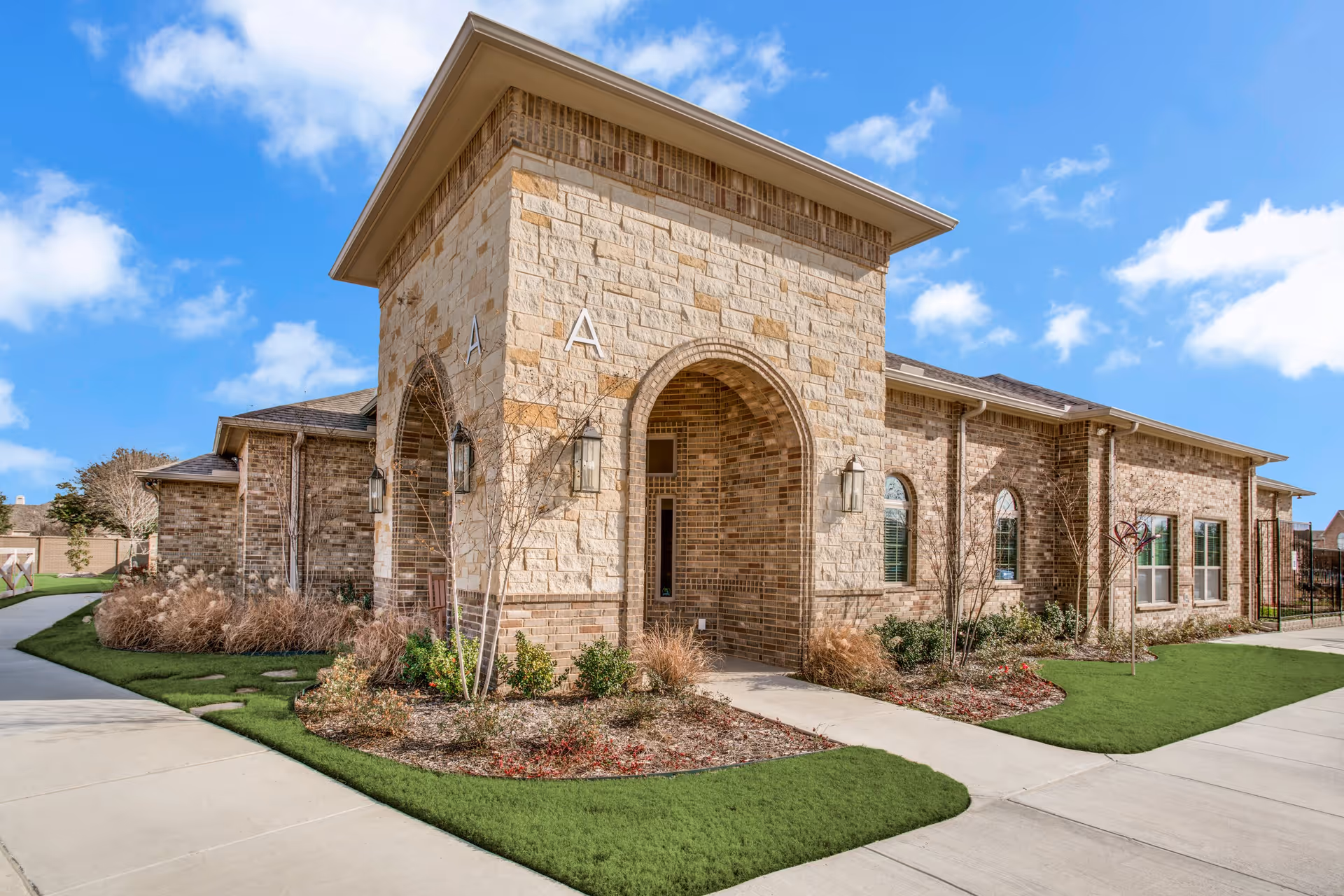 Exterior view of a single-story brick and stone building with arched entryways, landscaped garden beds, and a clear blue sky with some clouds.