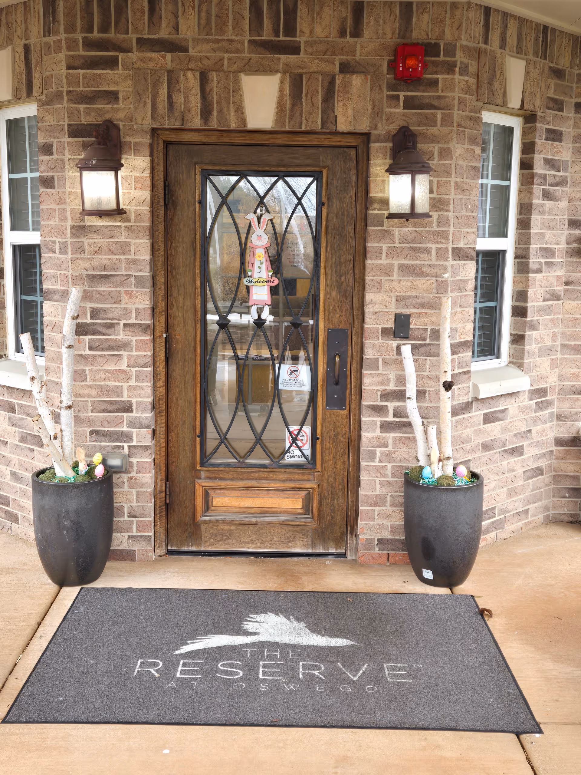 Brick building entrance with wooden door, decorative glass, lanterns, and mat reading The Reserve at Oswego.