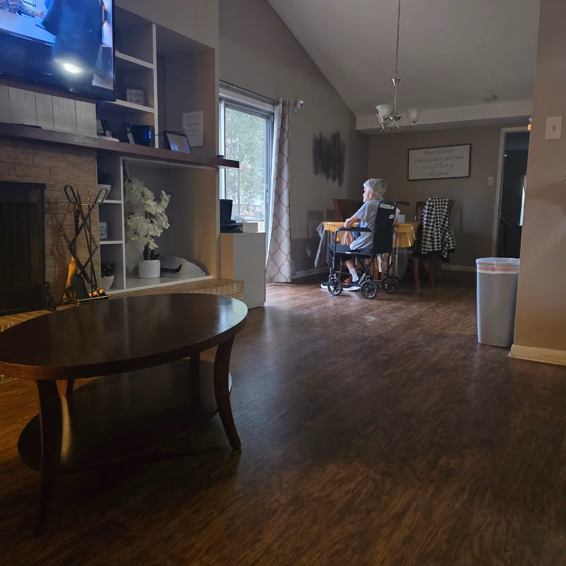 An elderly person in a wheelchair sits at a dining table in a cozy living room.