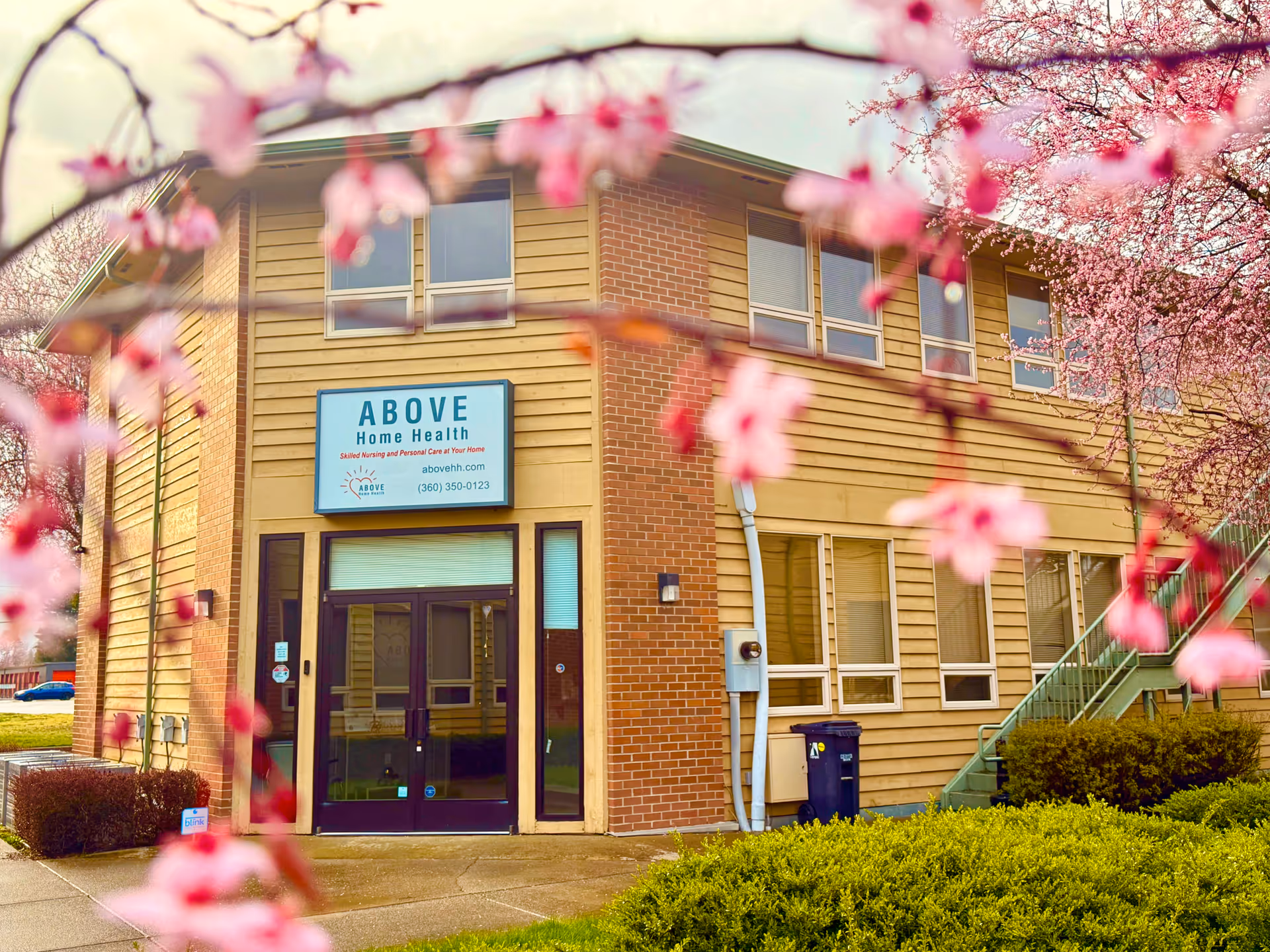 Two-story brick facility front with sign ABOVE Home Health framed by pink blossoms.