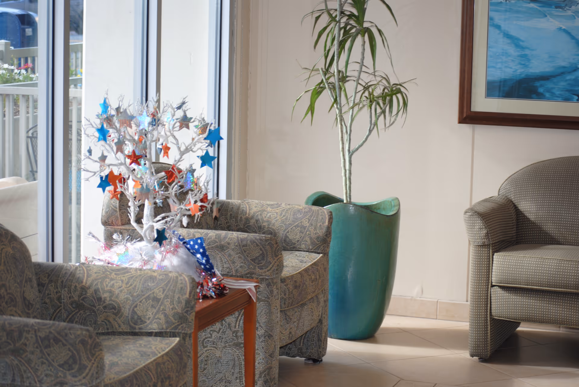 A cozy sitting area with patterned armchairs around a small wooden table. On the table is a decorative white tree adorned with red, white, and blue star ornaments. Behind the chairs is a large green plant in a tall green pot, and a framed picture hangs on the wall. Large windows let in natural light.