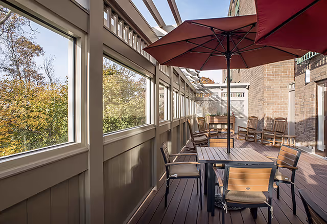 Outdoor patio area with wooden deck flooring, several wooden chairs, a table with a large red umbrella, and a brick building wall. The patio is enclosed with large windows and overlooks trees with autumn foliage.