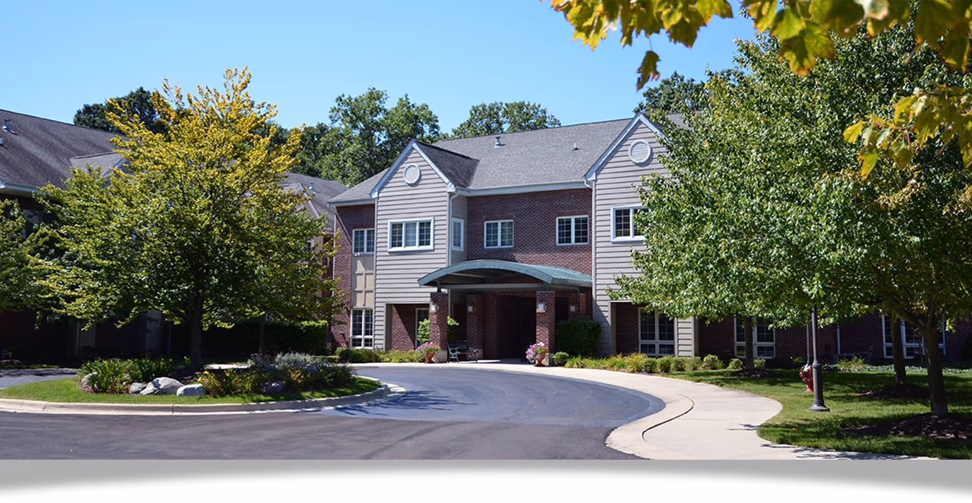 Front entrance of a two-story retirement community building with a covered porte-cochère, circular driveway, and landscaped trees.