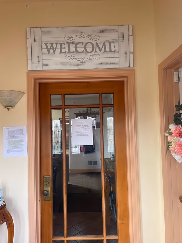 A wooden door with glass panels and a 'WELCOME' sign above it. There are papers taped to the wall and door, a wall-mounted light fixture on the left, and a floral decoration on the right side of the door frame.