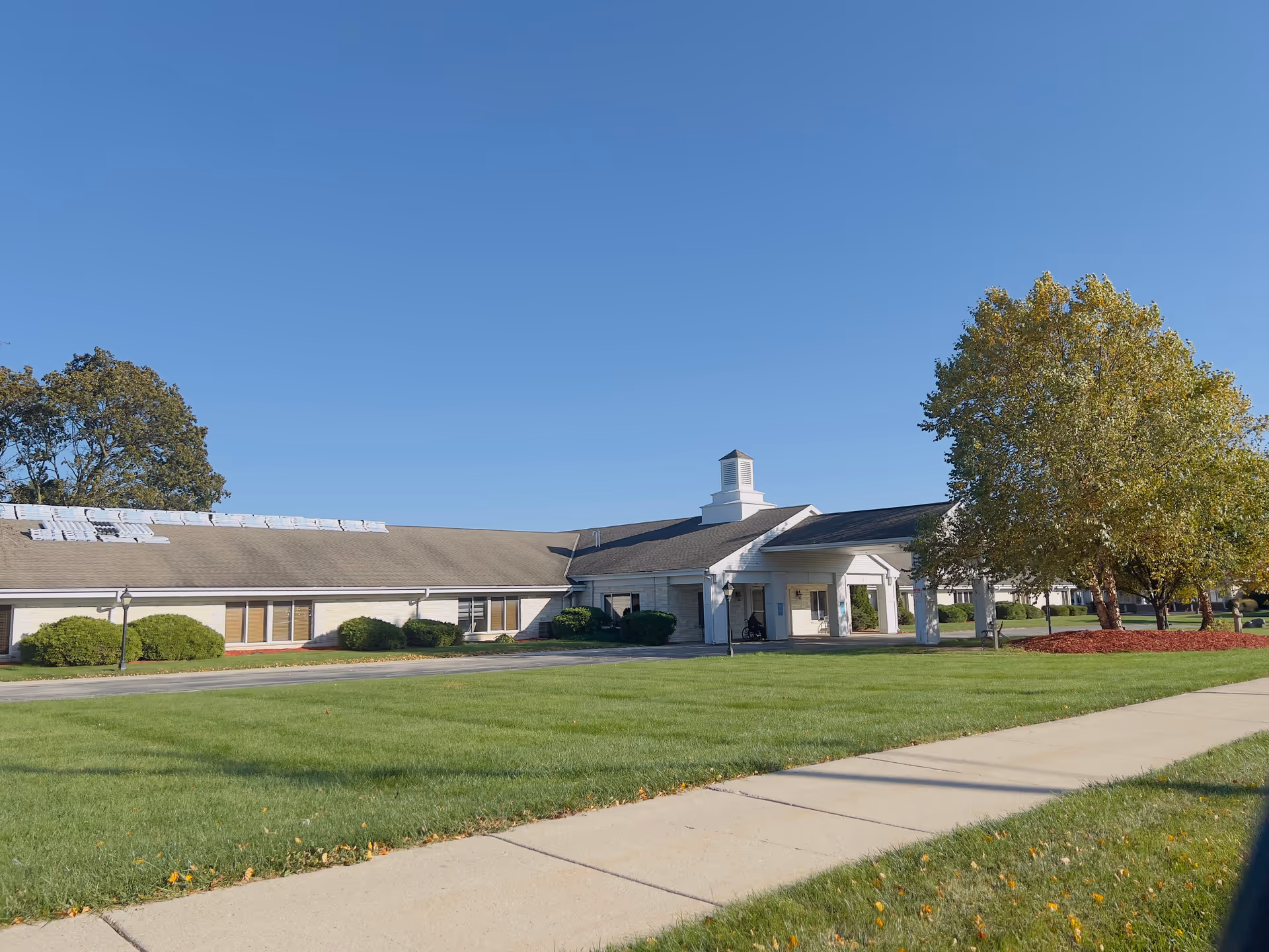 Single-story senior living building with a covered main entrance, wide lawn, and trees under a clear blue sky.