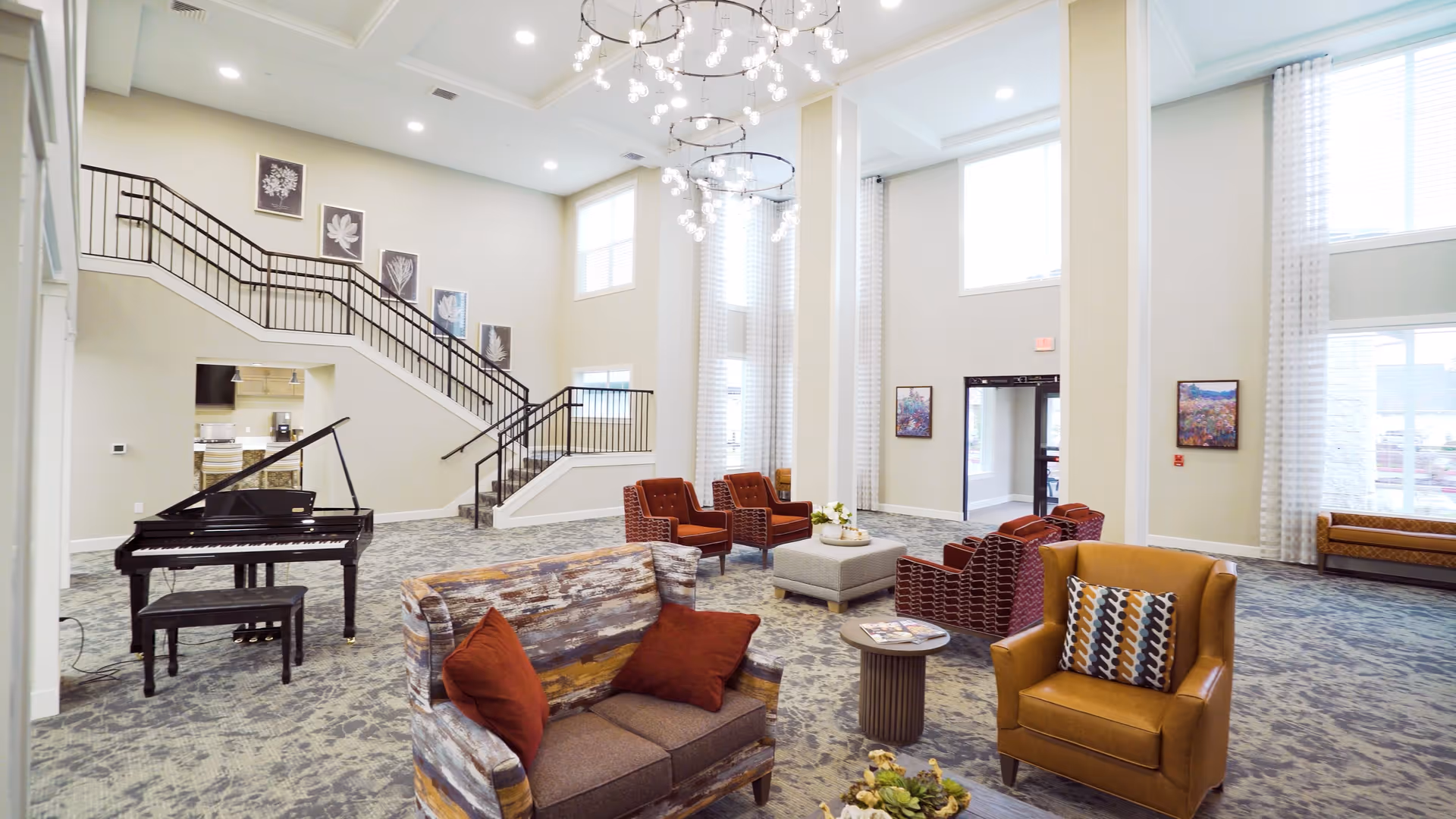Spacious and well-lit living room area with high ceilings and large windows. The room features a variety of seating options including a patterned sofa with red cushions, a mustard yellow armchair with a decorative pillow, and several red armchairs arranged around a central ottoman. A black grand piano with a matching bench is positioned near a staircase with black railings. The walls are decorated with framed artwork, and two modern chandeliers hang from the ceiling. The carpet has a subtle pattern, and there is a small round side table with magazines and a flower arrangement on the ottoman.