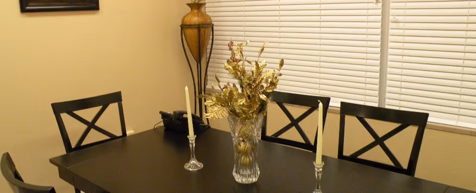 Dark wooden dining table with four chairs, a glass vase of flowers and two candlesticks in front of closed window blinds.