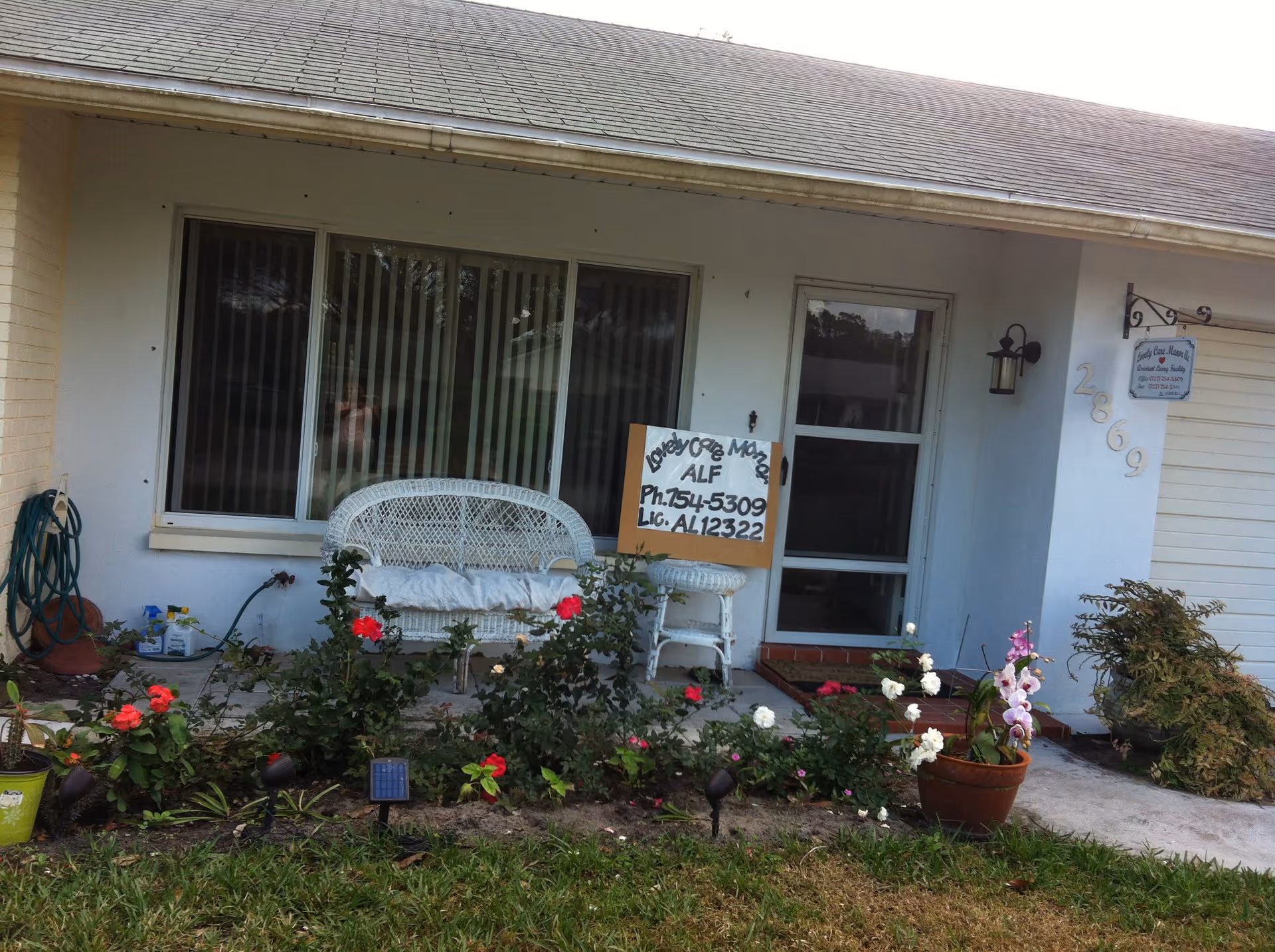 Front porch of Lovely Care Manor showing a white wicker bench, potted plants, and a handwritten sign by the entrance.