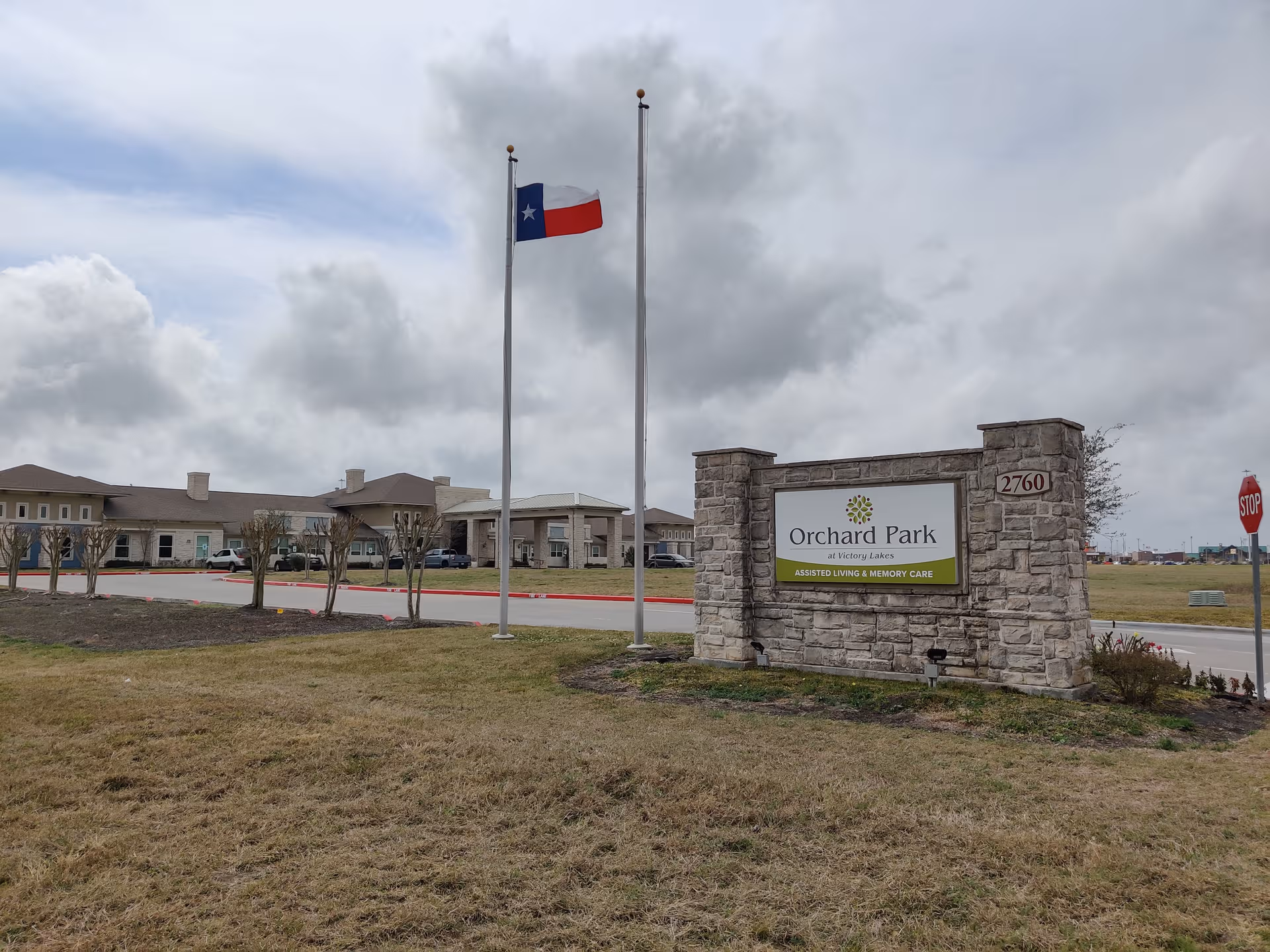 Stone entrance sign reads Orchard Park at Victory Lakes with flagpoles and a building in background.