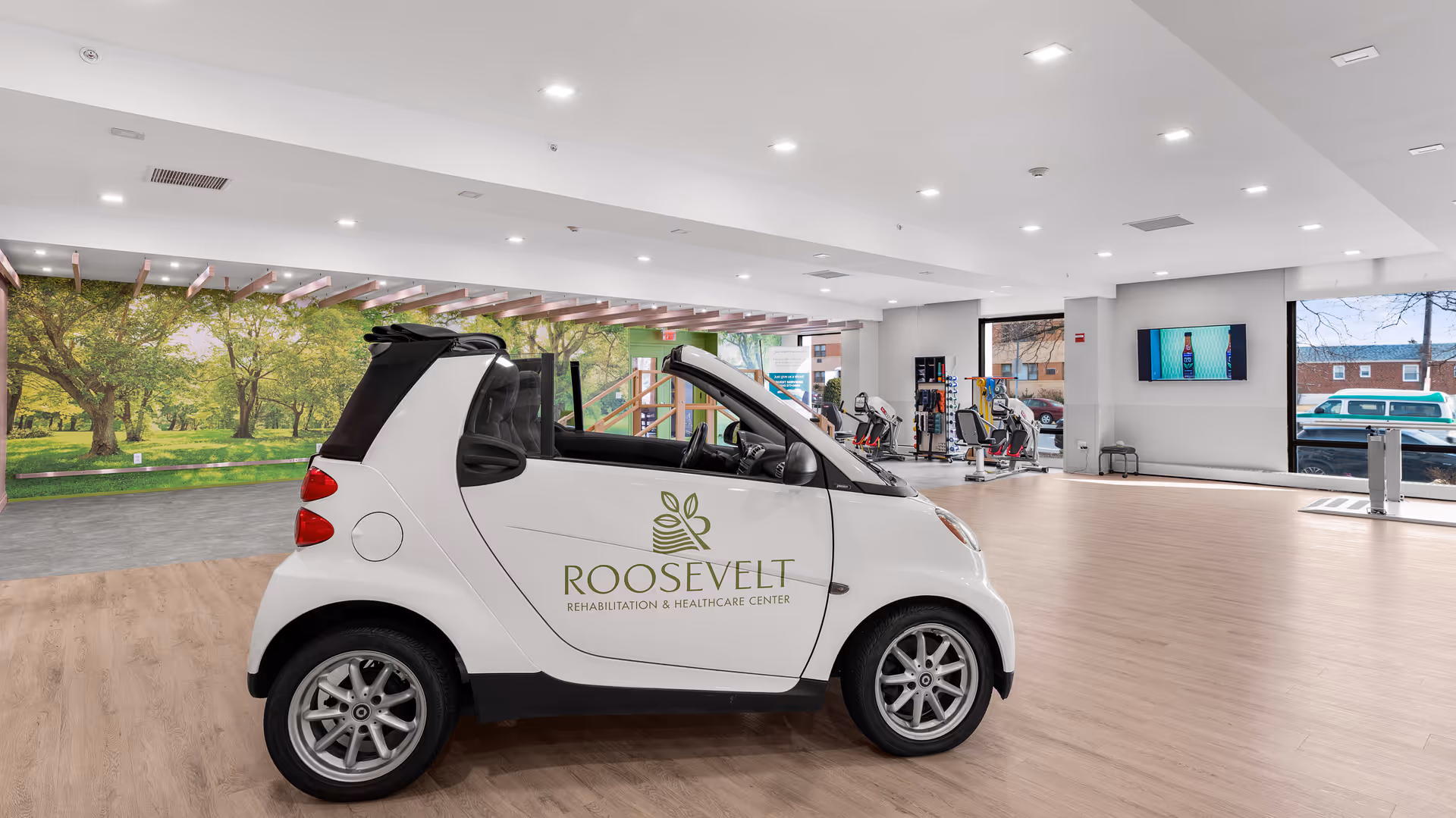 A small white convertible car with the logo and name 'Roosevelt Rehabilitation & Healthcare Center' on its side is parked inside a spacious, well-lit room with wooden flooring. The room has large windows showing an outdoor view, exercise equipment in the background, and a large wall mural depicting a green park with trees.