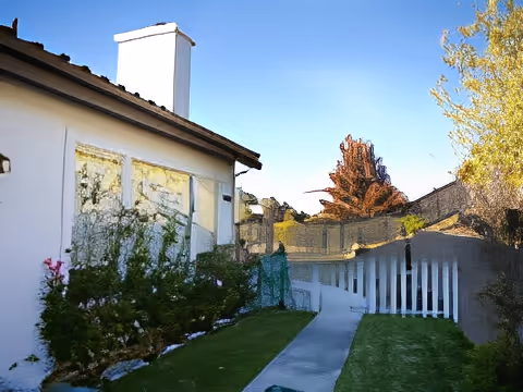 Outdoor view of a pathway leading alongside a white building with a chimney, surrounded by green bushes and grass, with trees and a clear blue sky in the background.