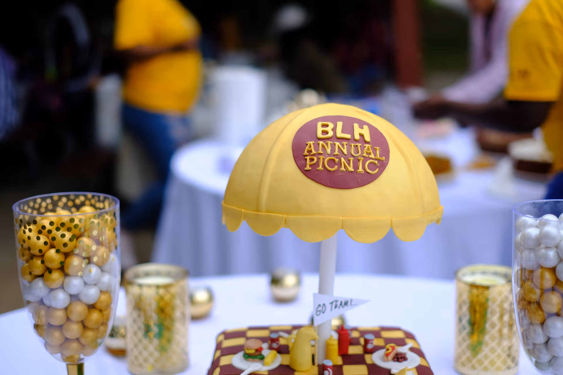 Outdoor picnic table with yellow BLH Annual Picnic umbrella centerpiece.