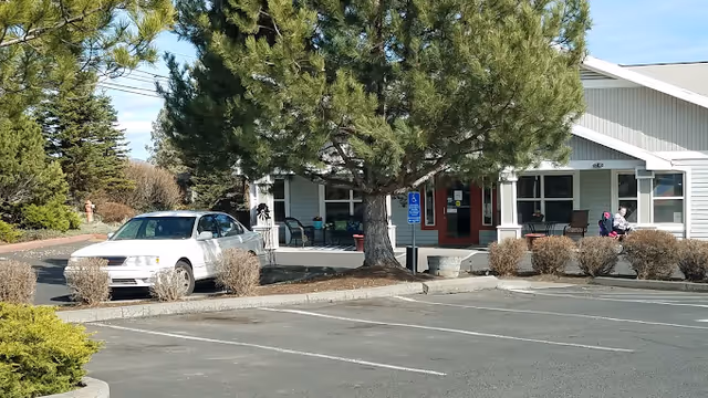 Exterior view of a single-story building with a covered entrance, surrounded by trees and bushes. A white car is parked near the entrance, and two people are sitting on chairs on the porch. The parking lot in front is mostly empty with marked parking spaces, including a handicapped parking sign near the entrance.