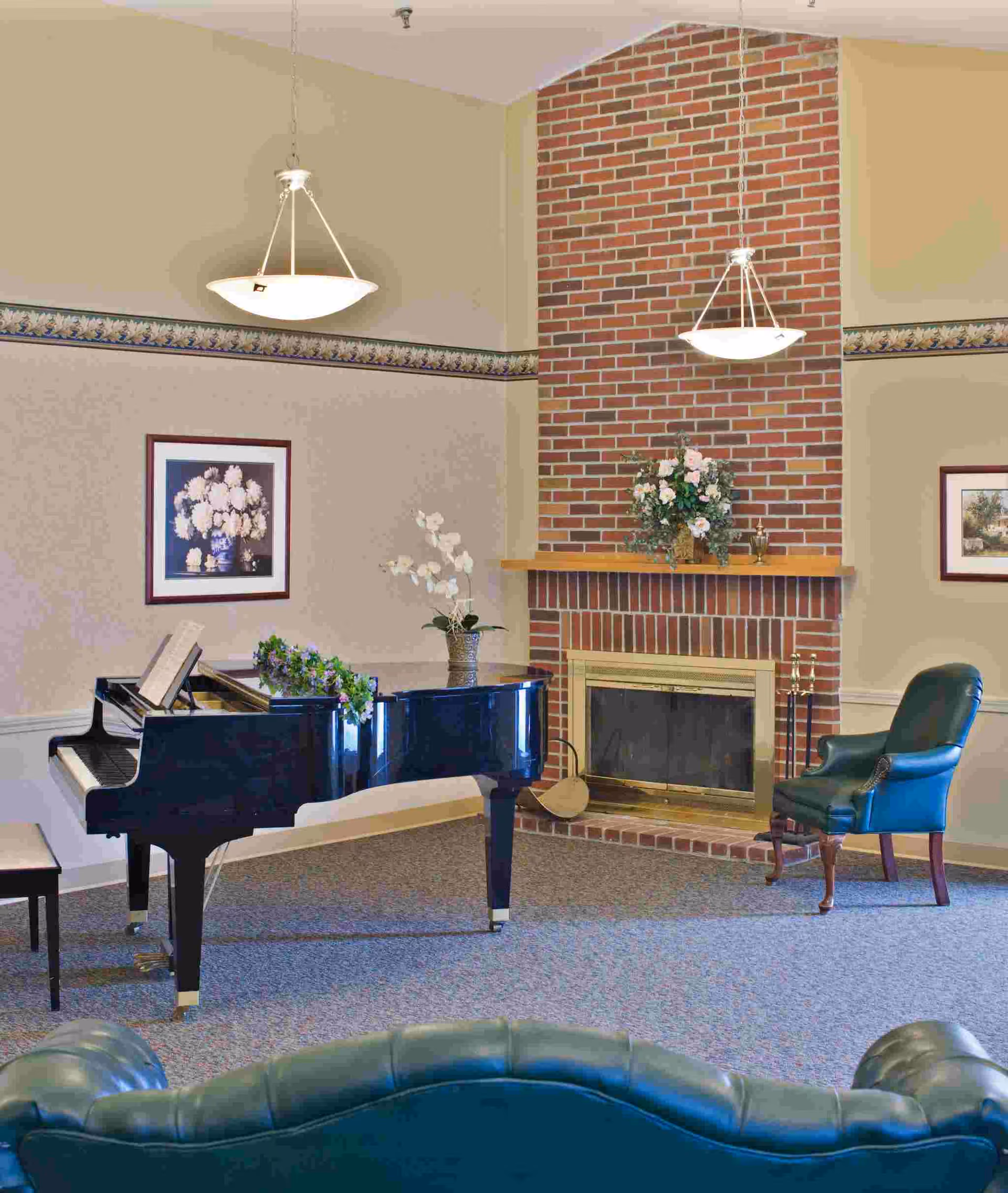 A communal lounge with a black grand piano, a brick fireplace topped with floral arrangements, and green leather seating.