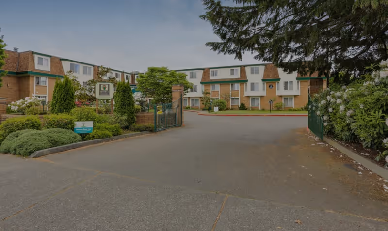 Gated driveway leading to a three-story brick senior living building surrounded by shrubs and trees.