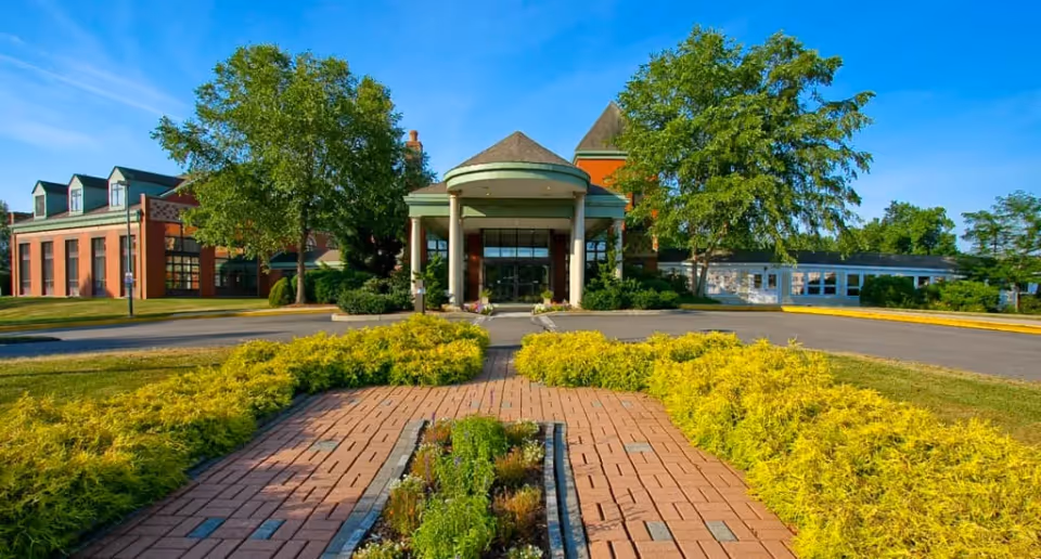 Front exterior view of a senior living facility named Evergreen Woods, featuring a circular driveway, a covered entrance with columns, well-maintained landscaping with green shrubs and trees, and a clear blue sky.