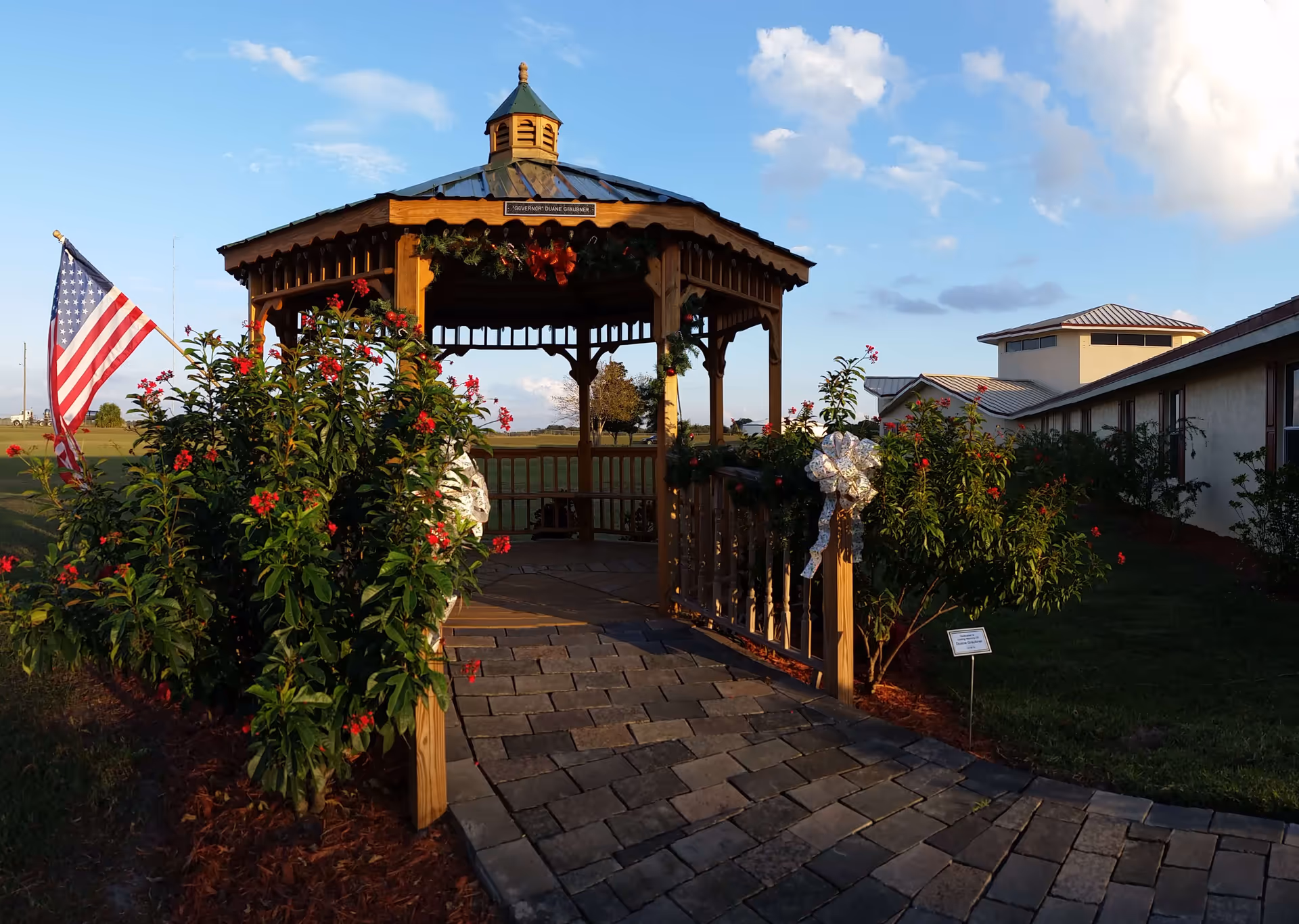 A wooden gazebo decorated with holiday garlands and bows, surrounded by green bushes with red flowers. An American flag is displayed on the left side. The gazebo is situated on a paved walkway with a building visible in the background under a partly cloudy sky.