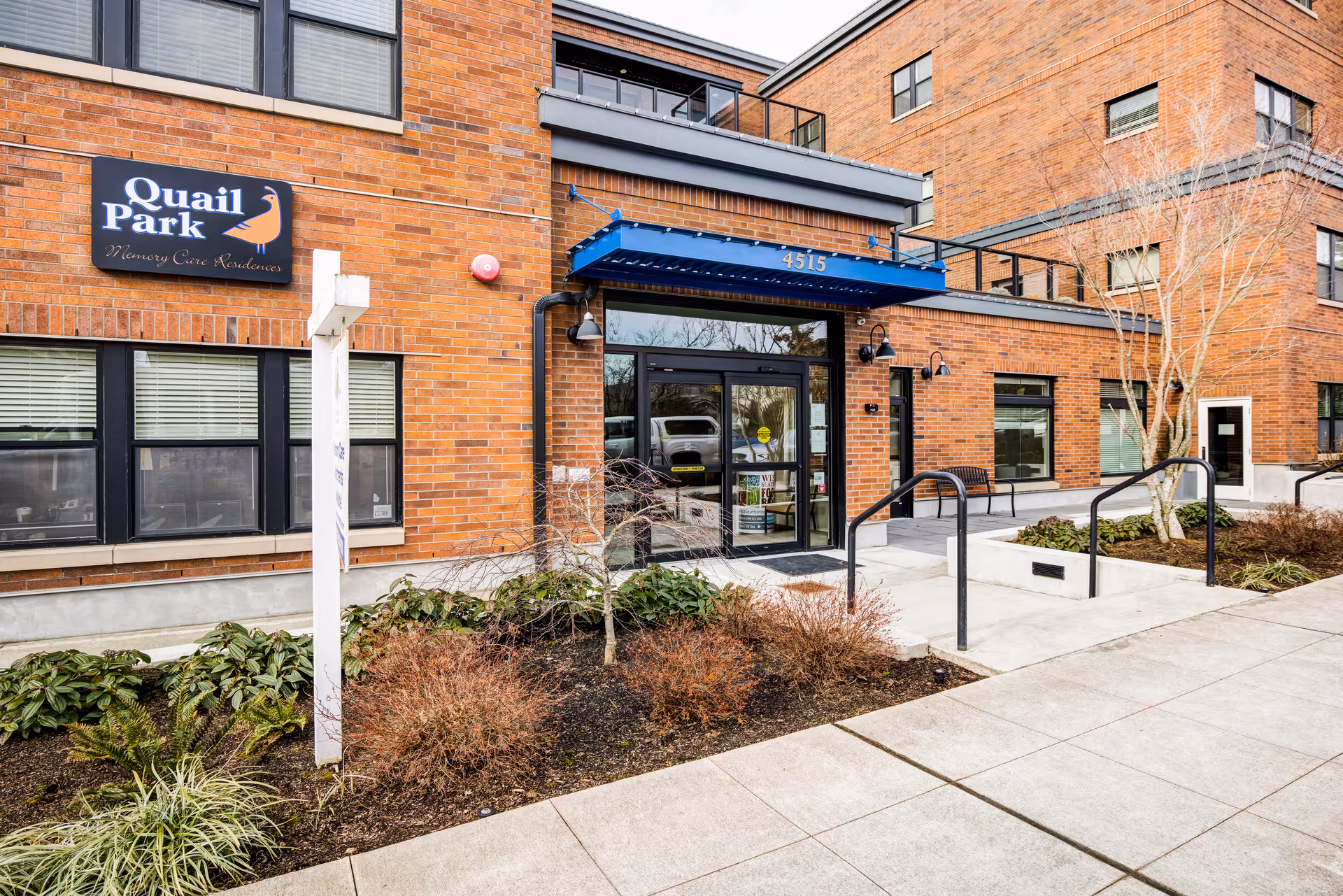Brick building entrance with blue awning and Quail Park sign, walkway and benches.
