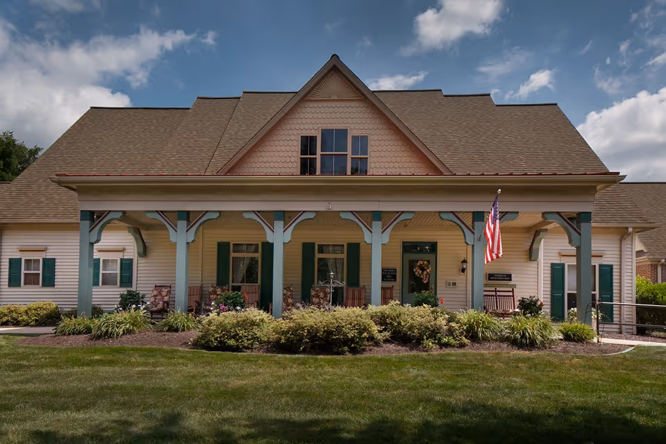 Cottage-style senior living front with a covered porch, greenery, and flag.