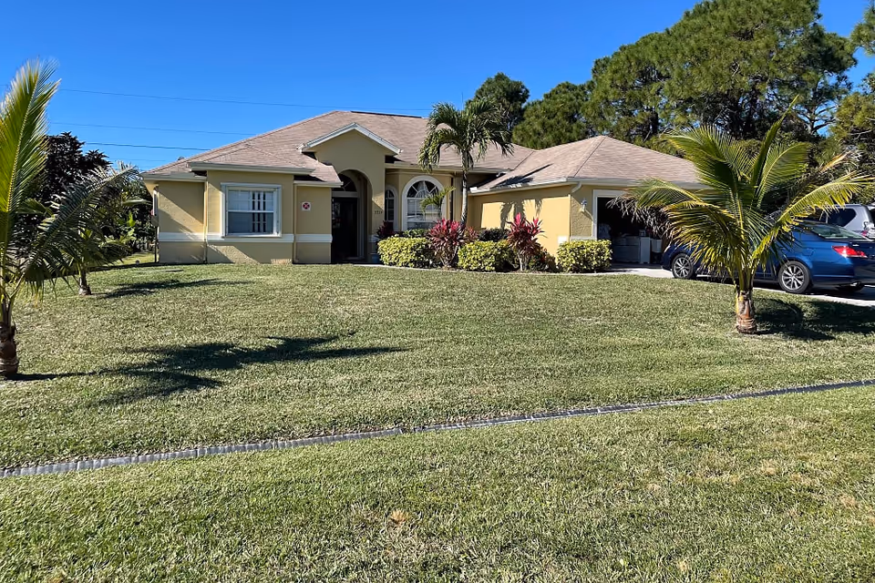 Single-story yellow house with a manicured lawn, palm trees, and a driveway with cars under a clear blue sky.