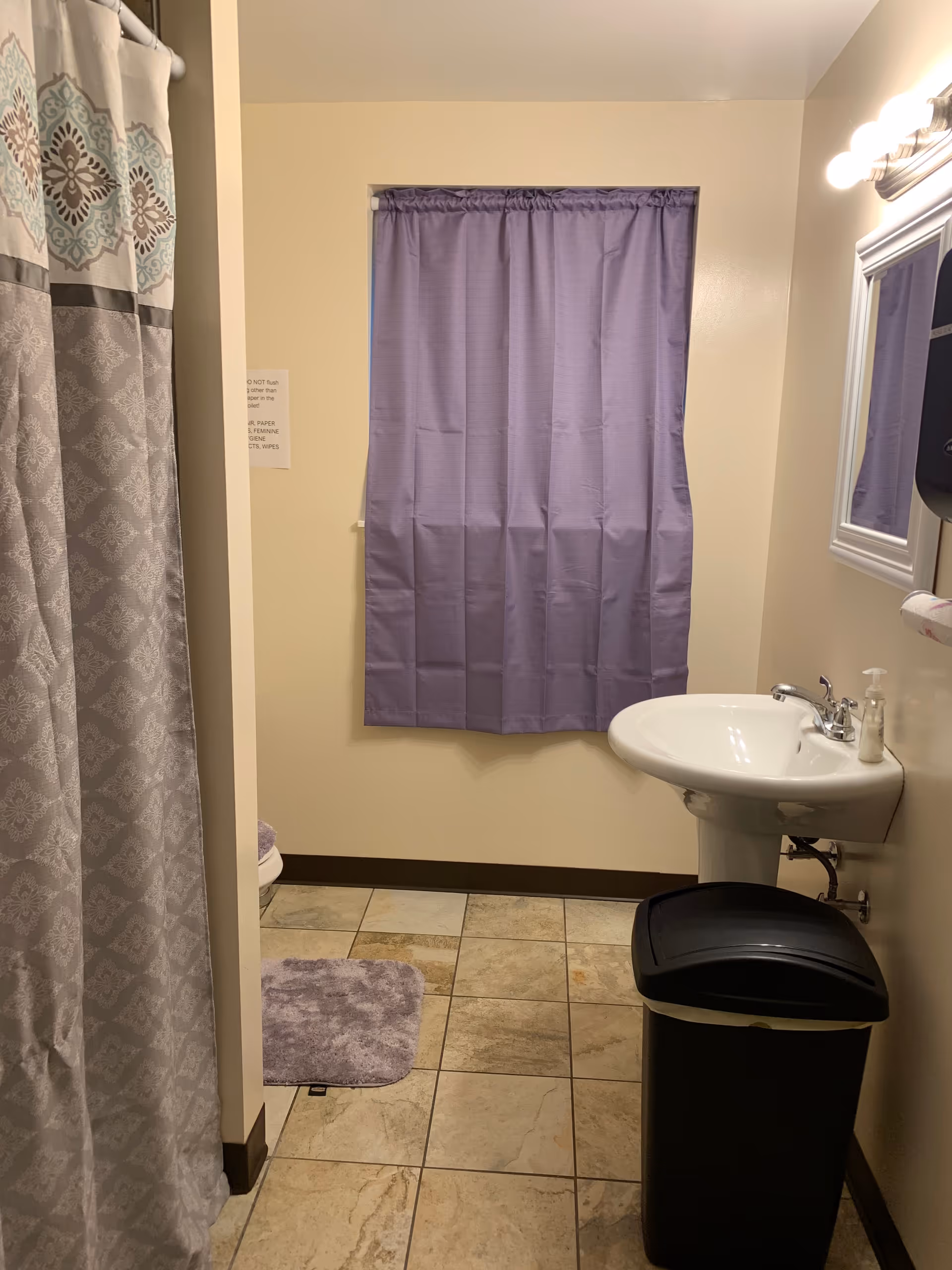 Small bathroom with purple curtain, shower, sink, mirror, and trash can.